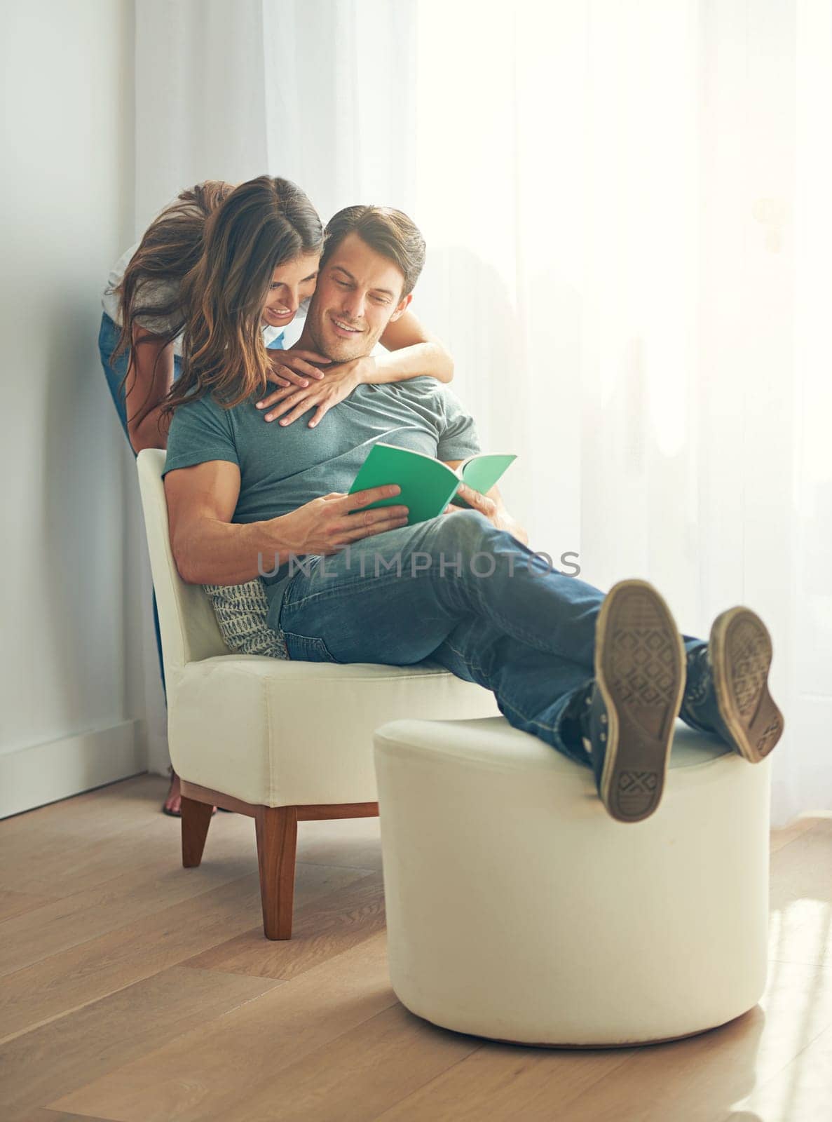 Couple, happy and book in home, love and chair to relax and minimalist interior for learning. Man, woman and romance while embrace, smile and reading for entertainment and together in living room.