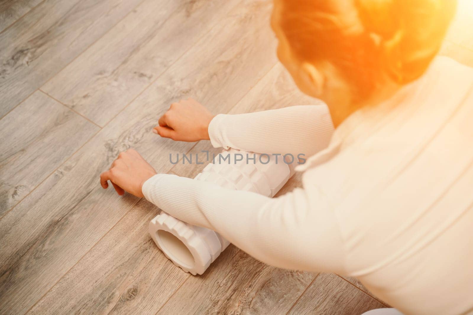 Adult athletic woman, in white bodysuit, performing fascia exercises on the floor - caucasian woman using a massage foam roller - a tool to relieve tension in the back and relieve muscle pain - the concept of physiotherapy and stretching training by Matiunina