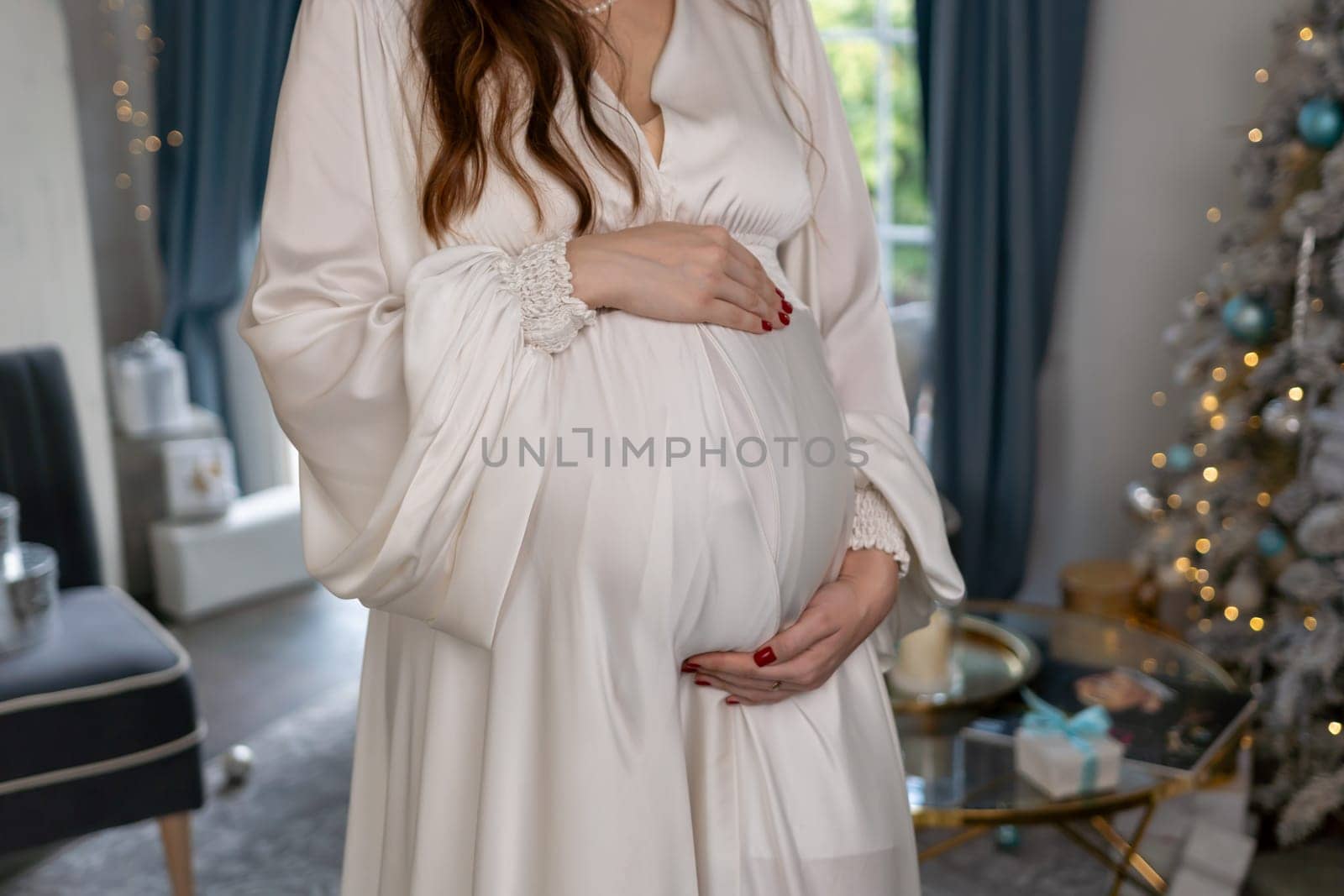 Pregnant woman holding a belly against Christmas tree with lights. family holiday concept. by Matiunina