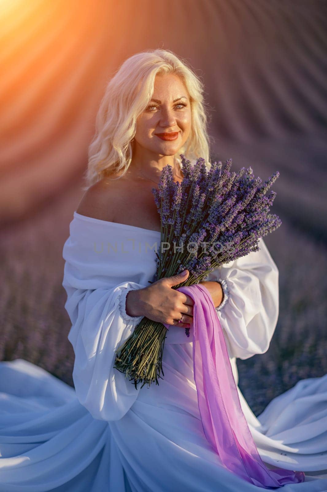 Blonde woman poses in lavender field at sunset. Happy woman in white dress holds lavender bouquet. Aromatherapy concept, lavender oil, photo session in lavender by Matiunina