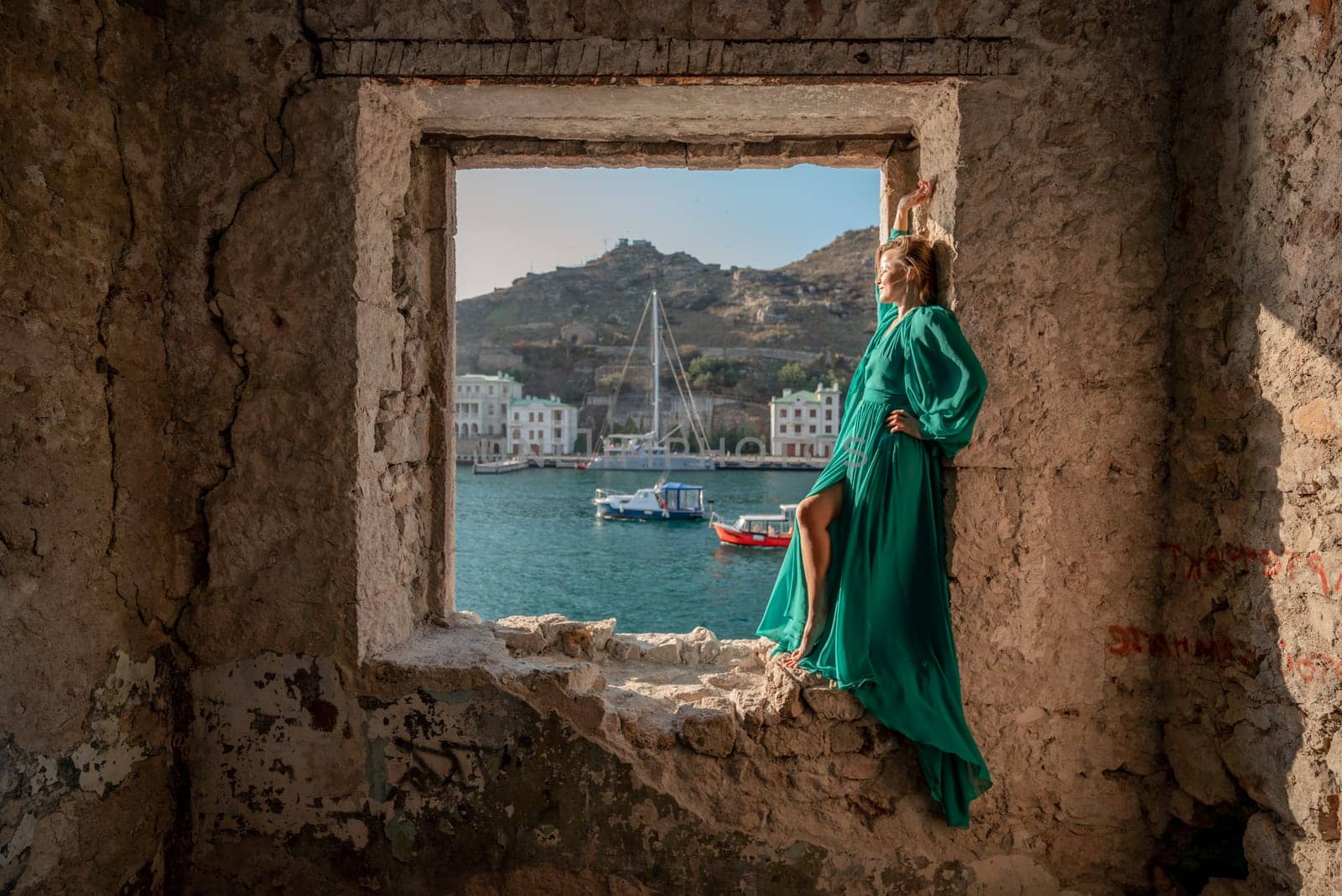 Rear view of a happy blonde woman in a long mint dress posing against the backdrop of the sea in an old building with columns. Girl in nature against the blue sky