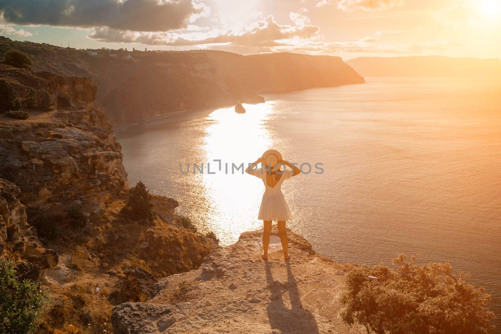 woman sea travel. photo happy woman with hat in white dress standing on the shore on a hill overlooking the sea. golden hour, silhouette of a woman at sunset on the mountain. by Matiunina