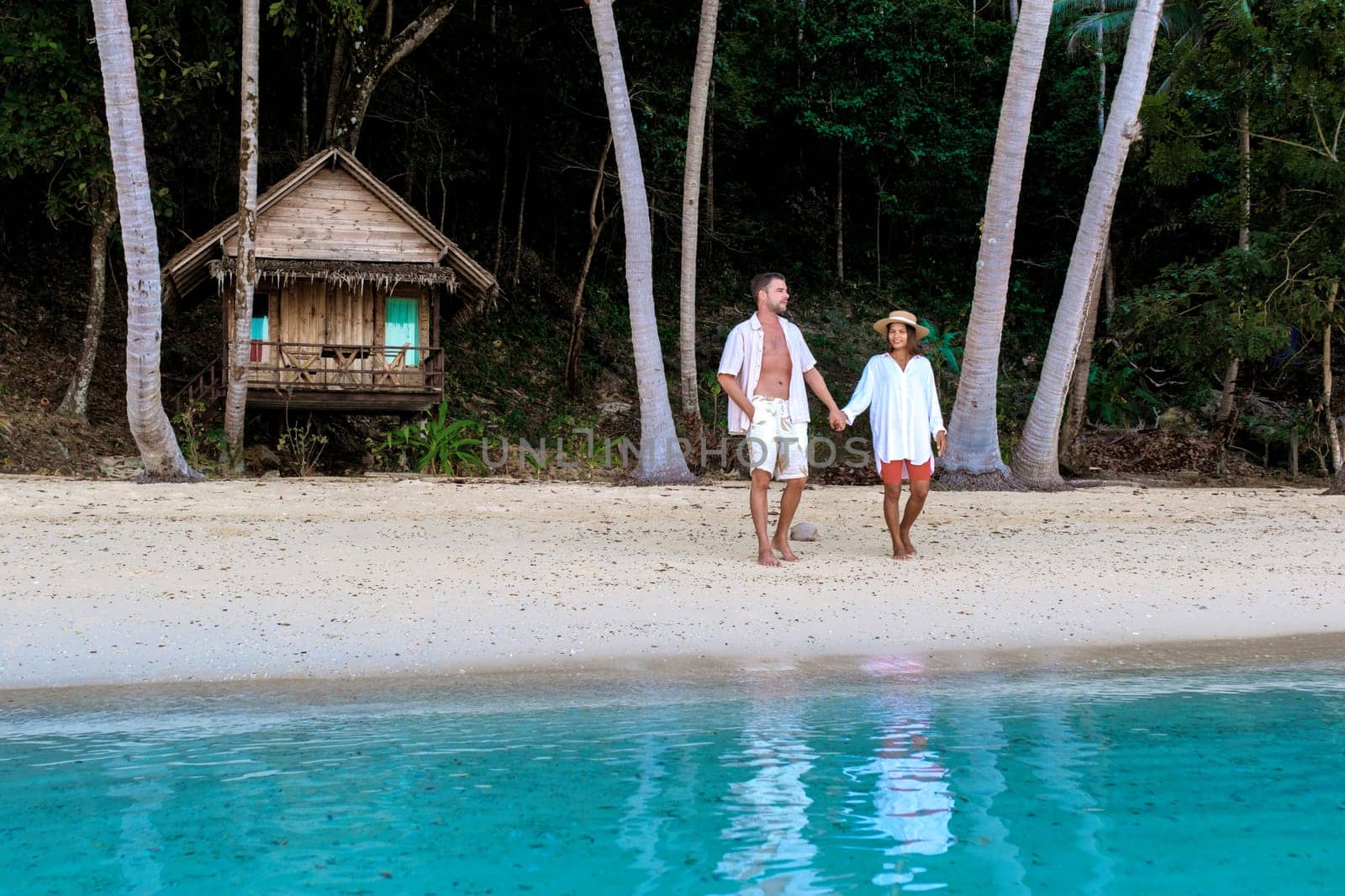 diverse couple of Asian woman and European man walking on the beach at sunset, on the background a wooden bamboo hut bungalow. a young couple of men and woman on a tropical Island in Thailand