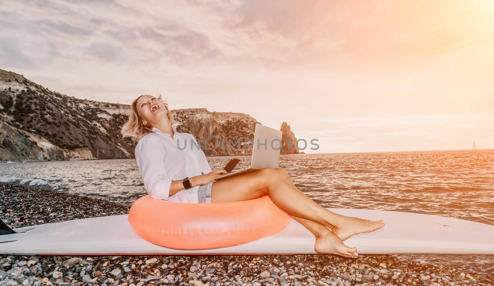 Successful business woman in yellow hat working on laptop by the sea. Pretty lady typing on computer at summer day outdoors. Freelance, travel and holidays concept.