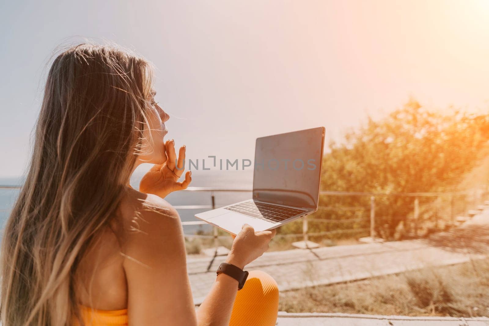 Woman laptop sea. Working remotely on seashore. Happy successful woman female freelancer working on laptop by the sea at sunset, makes a business transaction online. Freelance, remote work on vacation by panophotograph