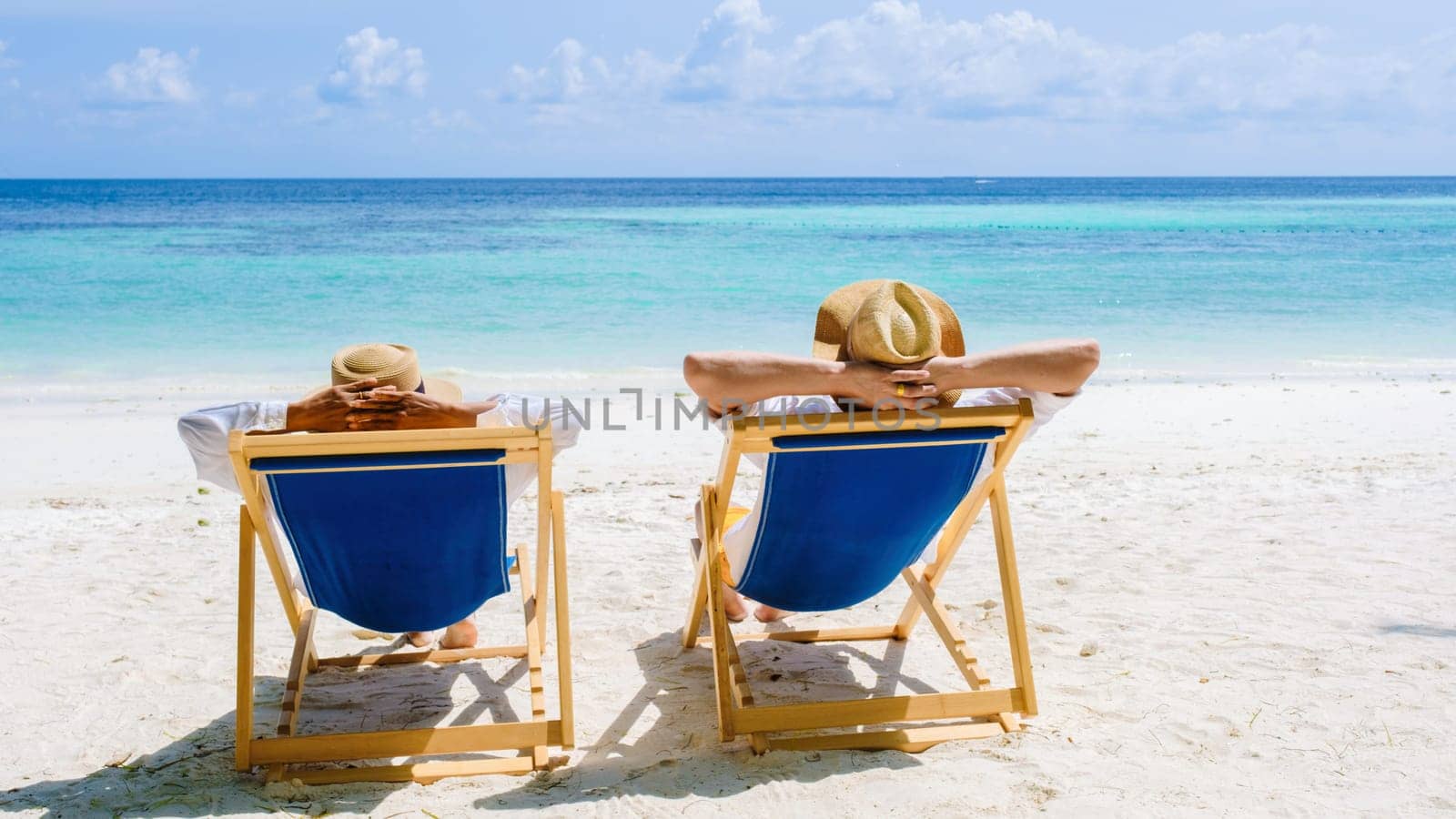 rear view of a couple of men and women sitting relaxing in beach chairs at Koh Lipe Island Southern Thailand, turqouse colored ocean and white sandy beach, diverse couple honeymoon or holiday concept