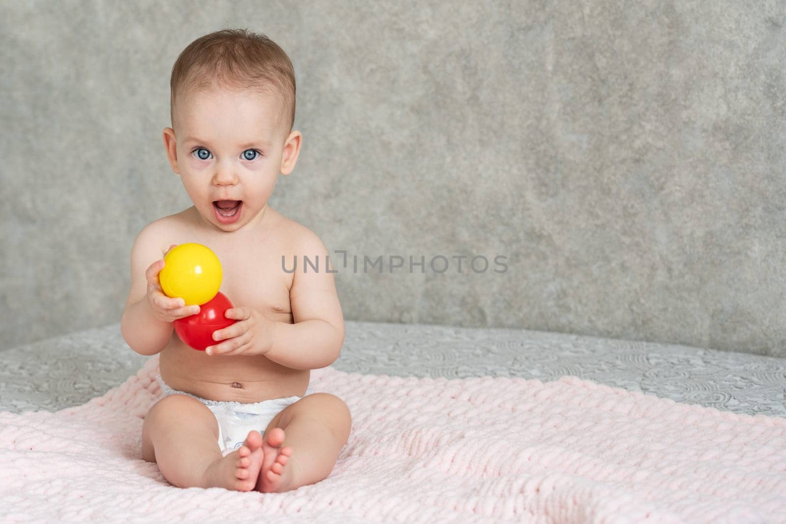 A happy toddler, wearing only a diaper, is sitting on a blanket, using their arm and thumb to hold