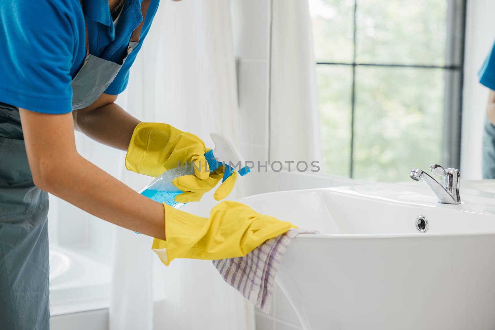 Maid with gloves scrubs limescale from bathroom sink and faucet applying spray detergent. Her commitment to housework ensures purity shining fixtures and hygiene. spray cleaner by Sorapop