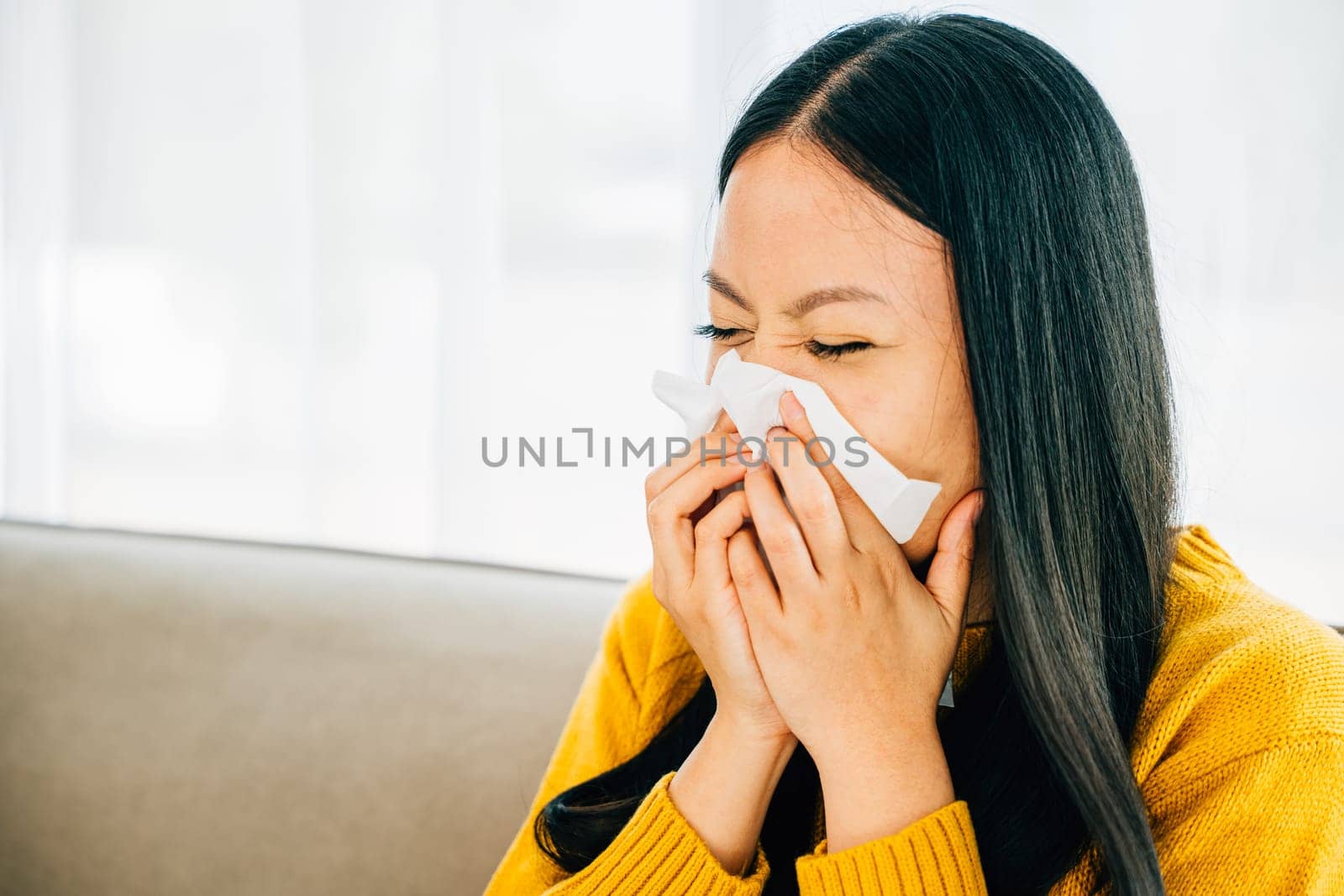 Portrait of an Asian woman in the living room holding a tissue dealing with cold symptoms like cough and sneezing. A patient at home managing flu. Snot and handkerchief in hand.
