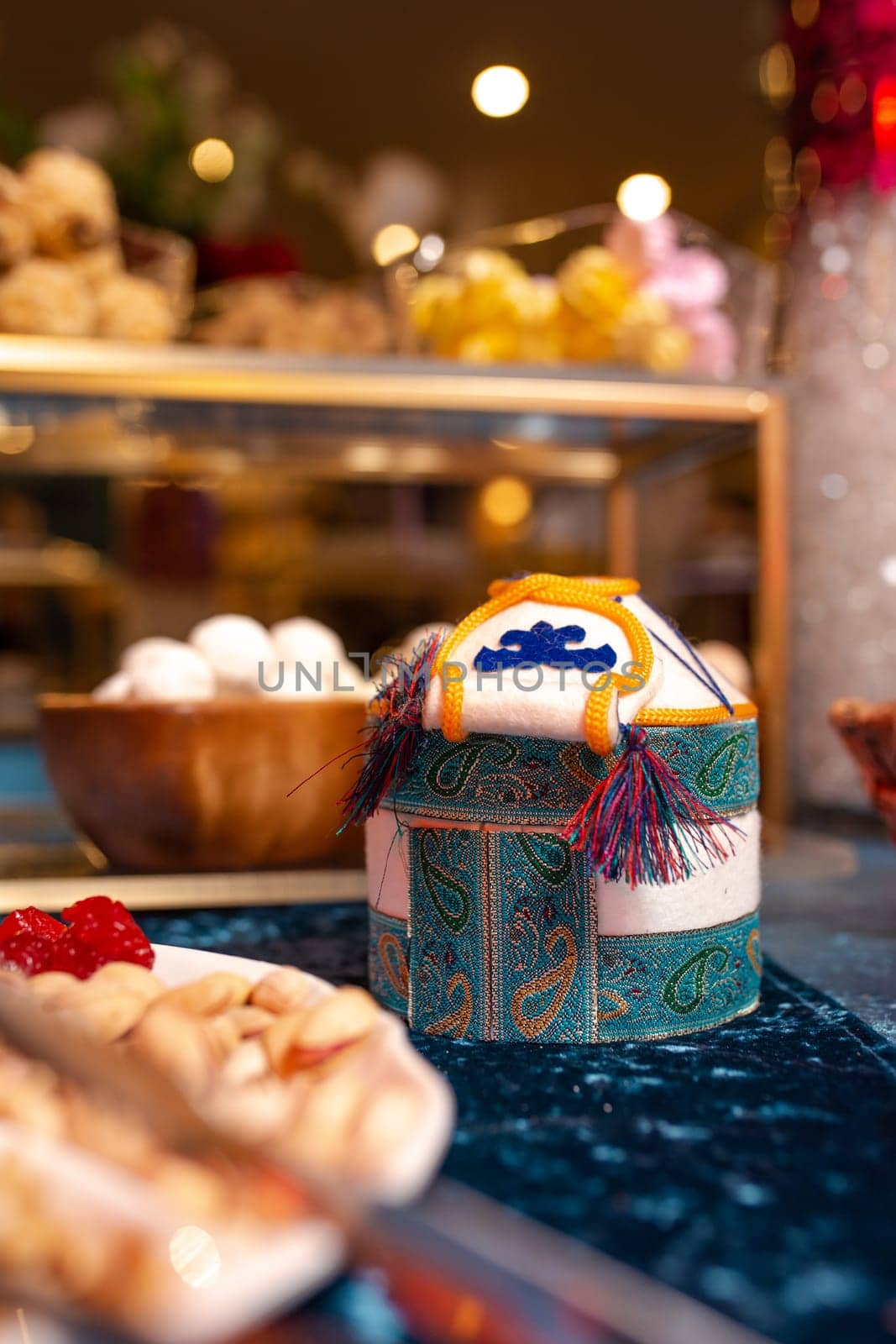 Yurt-shaped container with intricate patterns sits on a blue tablecloth, surrounded by an assortment of sweets and nuts. The background features a blurred buffet setting.