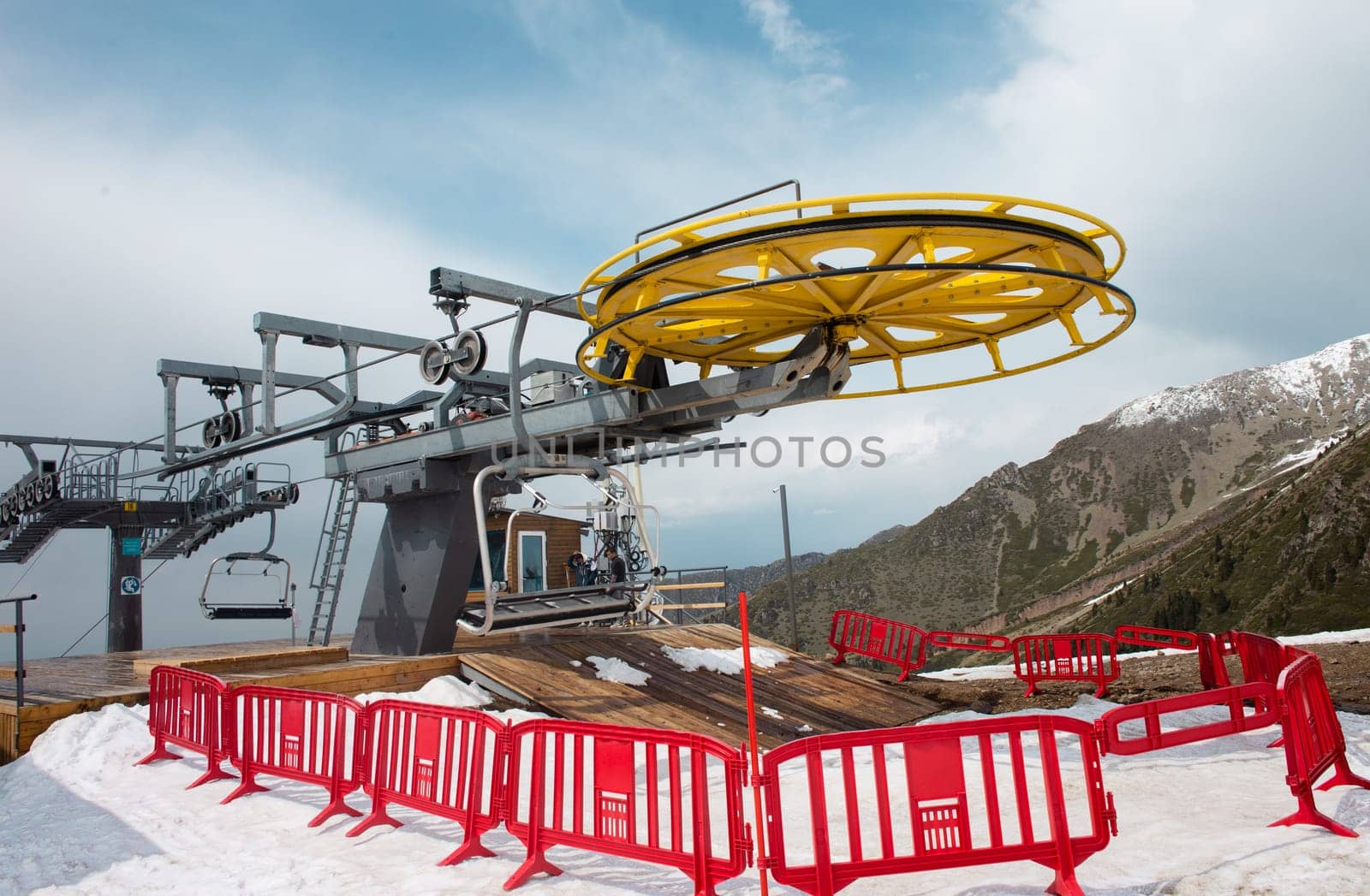 A yellow and gray ski lift wheel and tower with a blue sky and snow-capped mountains in the background. The ski lift is surrounded by red and white striped safety barriers.