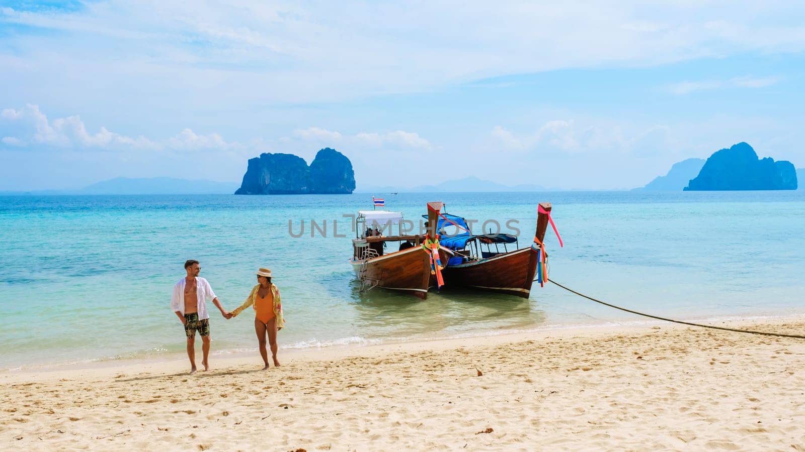 a couple of men and woman walking on the beach with longtail boats in the ocean of Koh Ngai island soft white sand, and a turqouse colored ocean in Thailand, diverse couple swim in the ocean