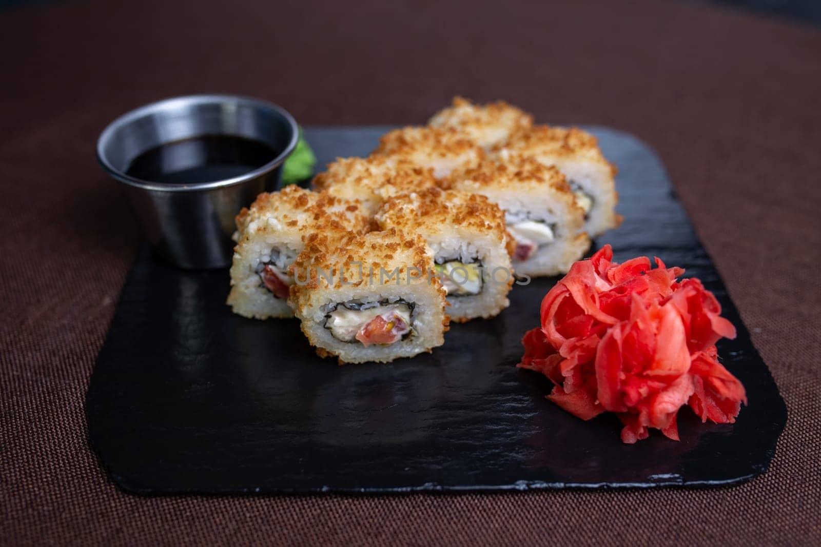 Deep-fried sushi roll on black stone plate with soy sauce, wasabi. Made with rice, seaweed, vegetables, shrimp tempura.