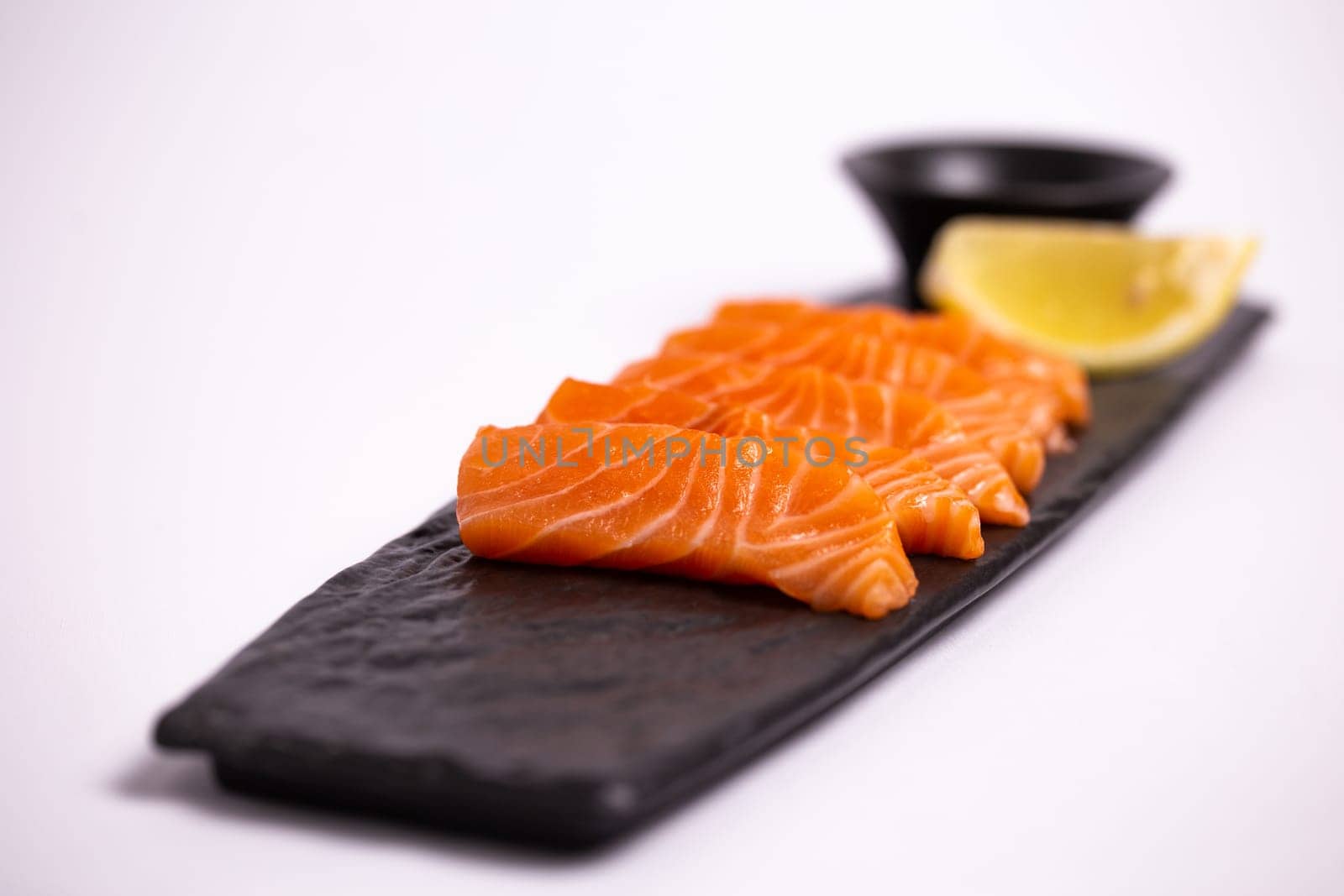 Top view of a delicious and healthy meal of fresh salmon fillet sliced into thin strips and served on a black stone plate with soy sauce and a lemon wedge. The plate is isolated on a white background.