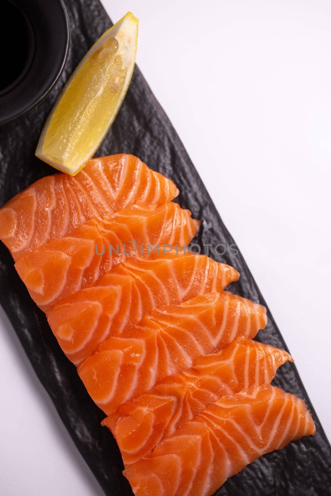 A close-up of a fresh salmon fillet garnished with lemon wedges and soy sauce on a black stone plate. This appetizing seafood dish is isolated on a white background, perfect for a healthy meal.