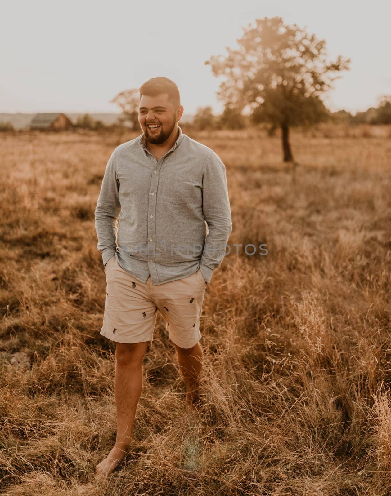 young caucasian black-haired overweight man with beard smiling cheerfully in summer cotton clothes and shorts outdoor in sunny weather. men fashion collection. open spaces meadow flooded sunlight tree