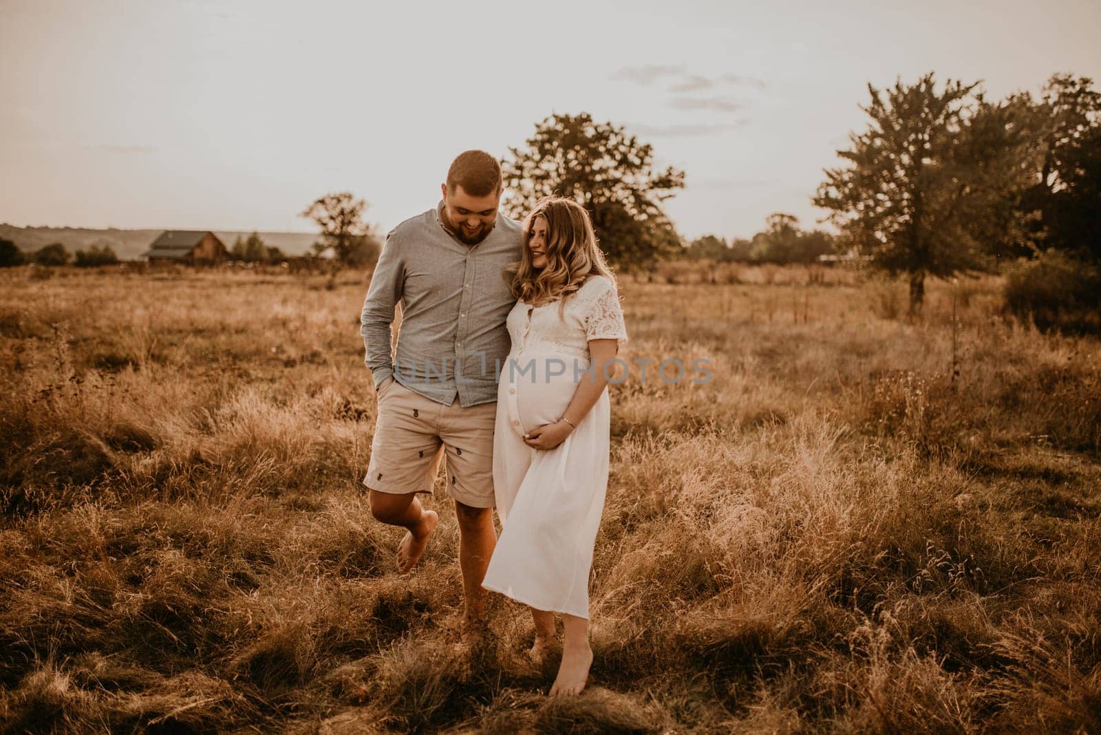 Happy family pregnant caucasian blonde woman with moles on face in white cotton dress walks with husband meadow summer. man in light natural clothes and shorts holds hand wife. trees in sunbeams
