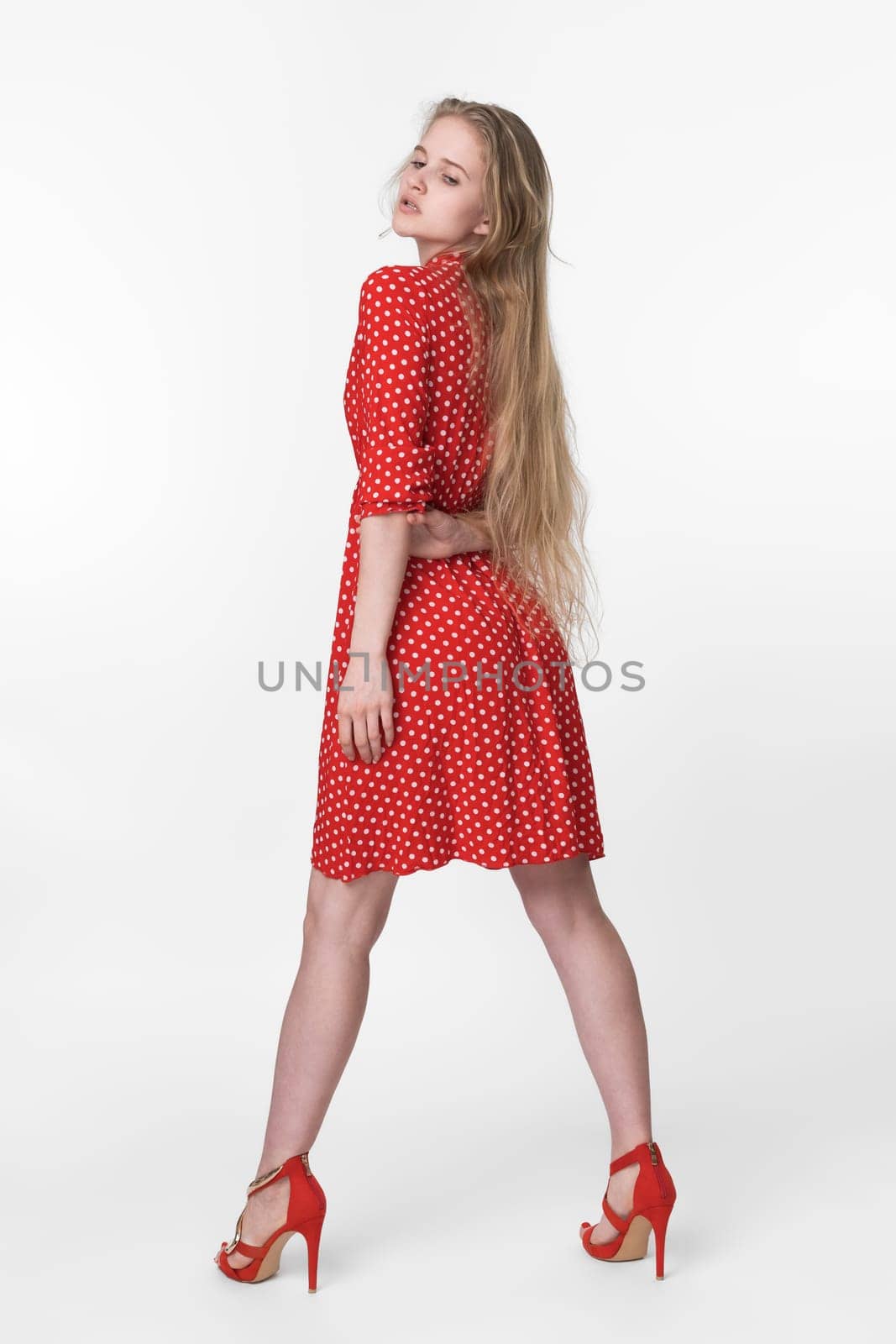 Stylish blonde woman with long hair dressed in summer red polka dot dress, red shoes standing in full length and poses. European female 21 years old looking down. Studio shot on white background