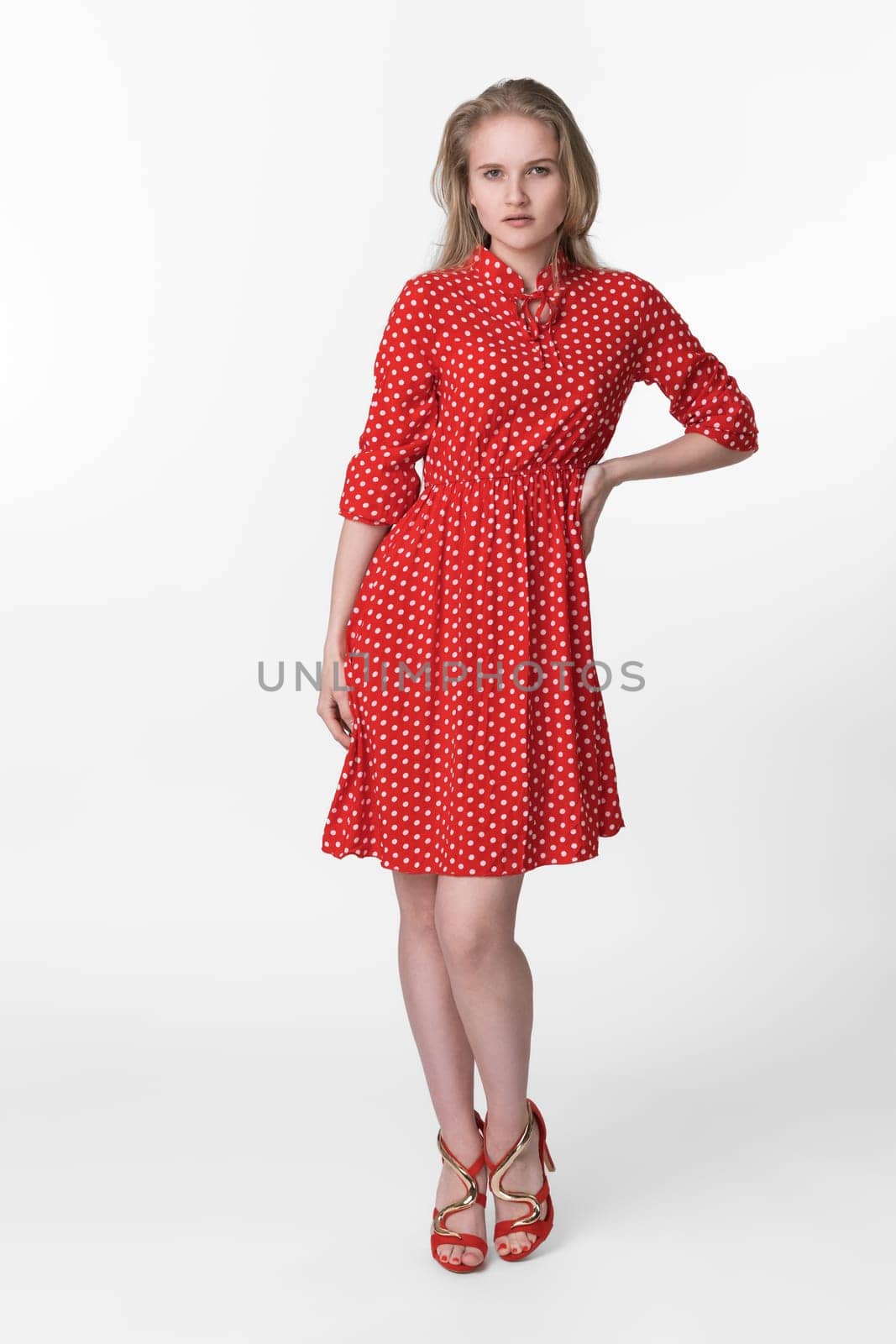 Young blonde woman with long hair dressed in summer red polka dot dress, red shoes standing in full length and poses on white background. Caucasian female 21 years old looking at camera. Studio shot