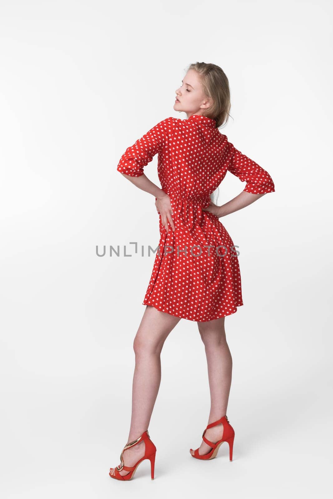 Cheerful blonde woman with closed eyes dressed in summer red polka dot dress, red shoes standing in full length and poses. Bossy European female 21 years old. Studio shot on white background