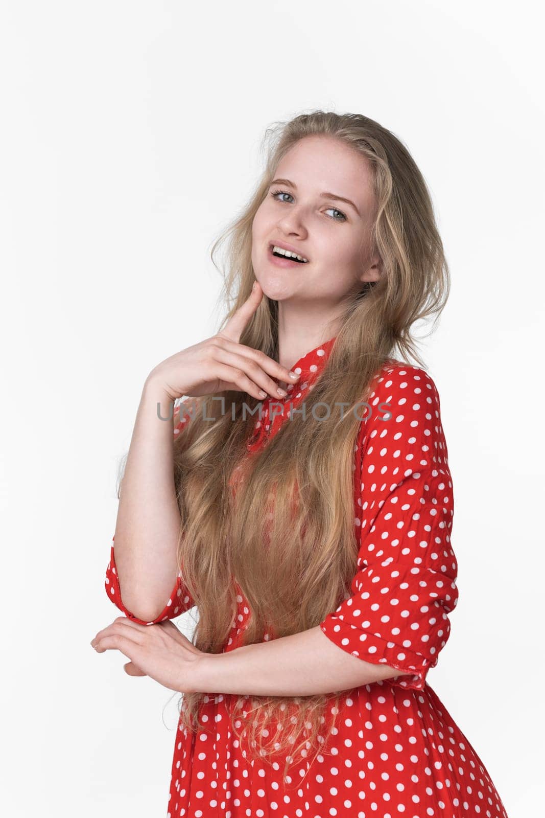 Playful young blonde female with long hair dressed in summer red polka dot dress poses on white background. Smiling Caucasian model 21 years old looking at camera. Studio shot, front view, waist up