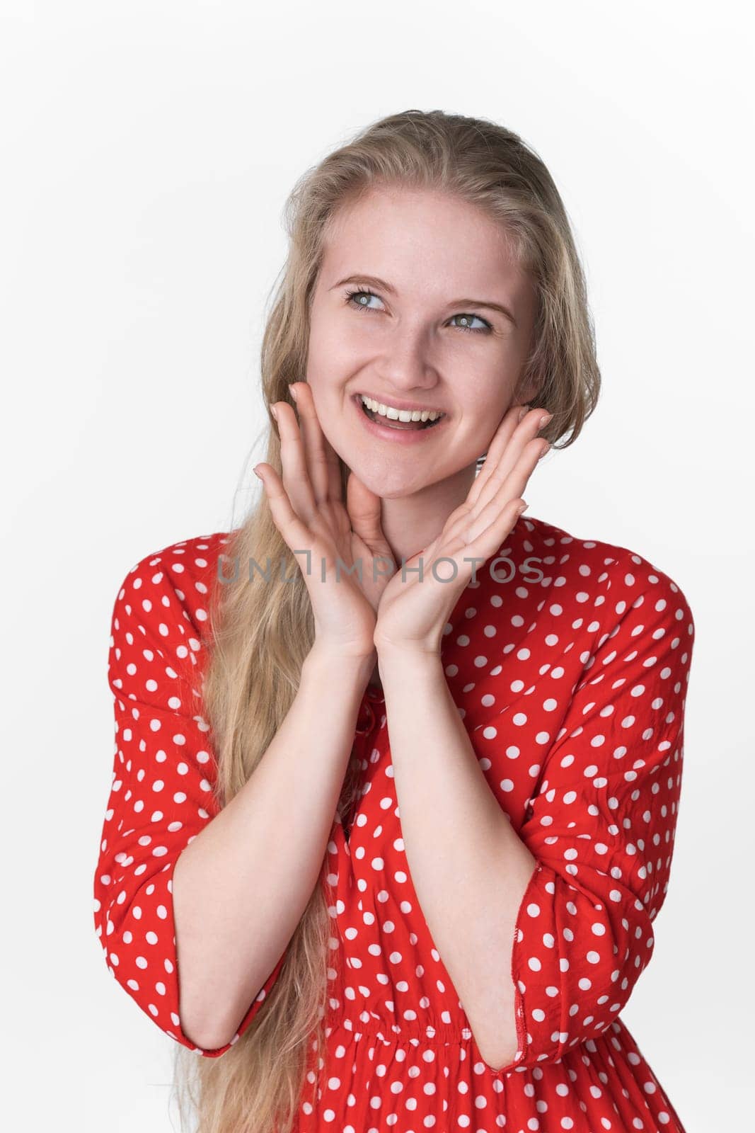 Portrait of blonde young adult woman with positive emotions on her face folded palm under chin and looking away. Playful 21 years old model in red polka dot dress. Studio shot on white background