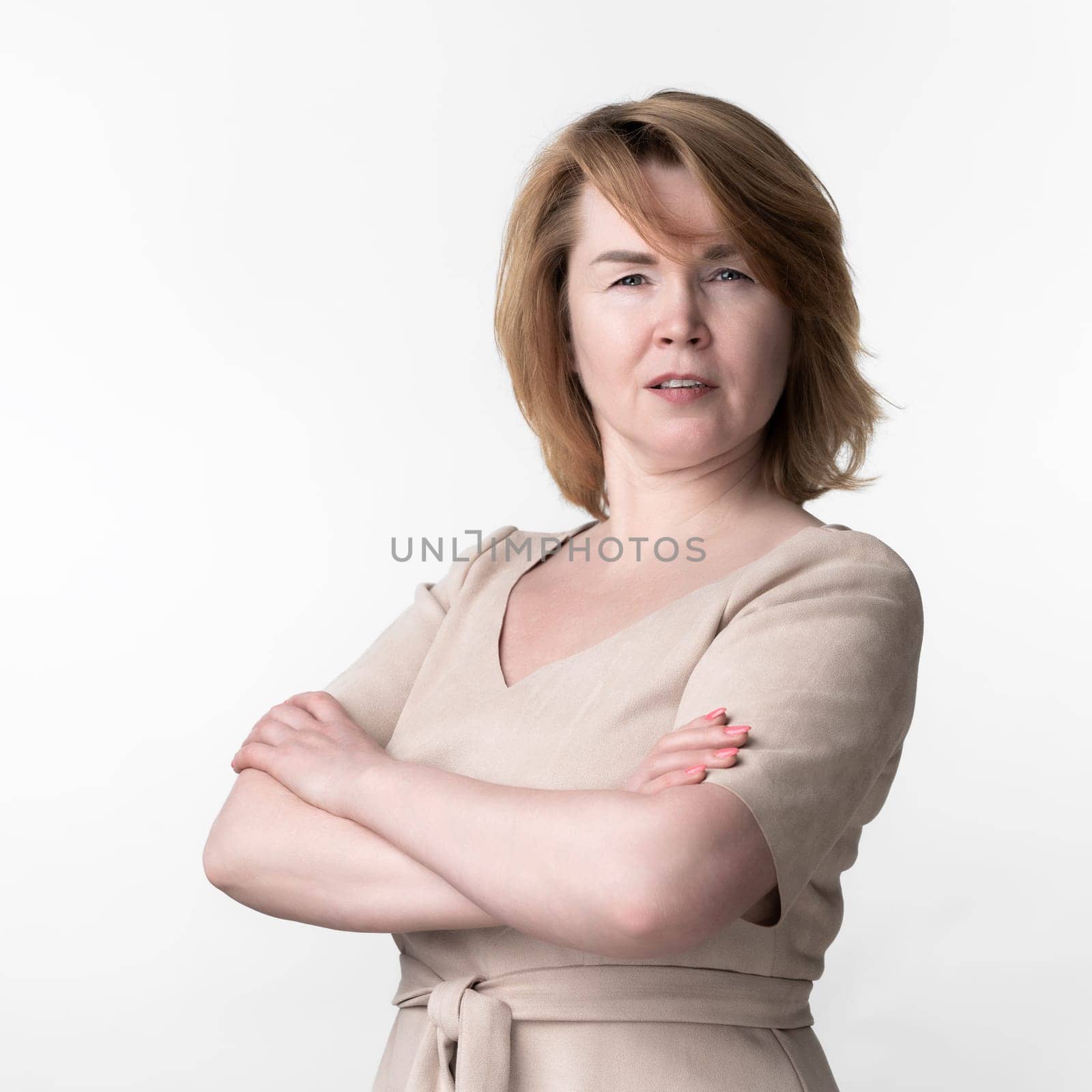 Portrait of serious woman 49 year old in beige dress with short sleeves and belt tied in front looking at camera carefully, with arms crossed on chest, standing on white background. Part of series