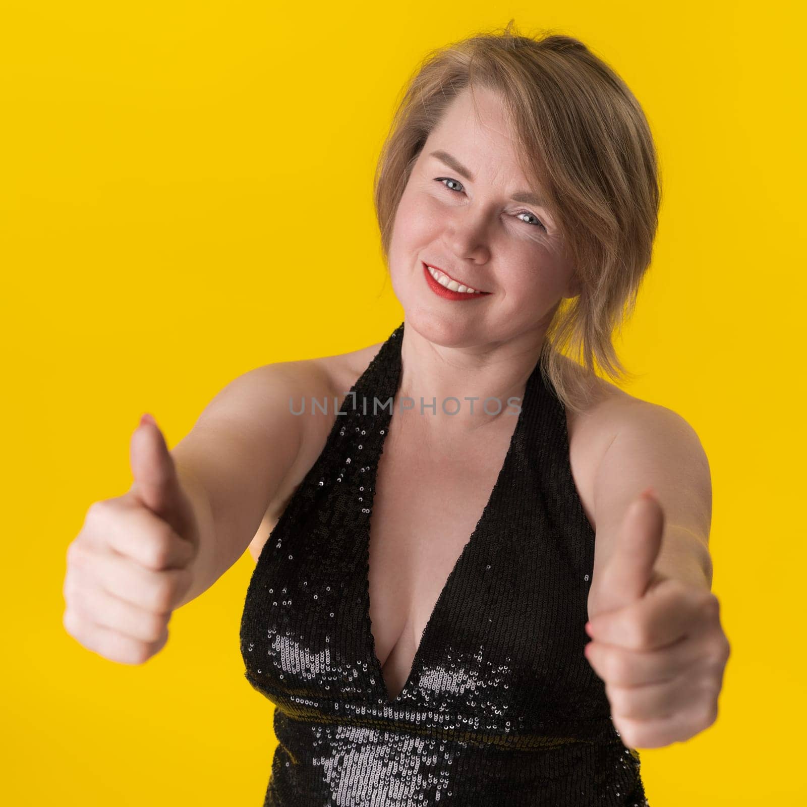 Portrait of curvy mature blonde woman in black evening gown with spangles and neckline on bright yellow background. Female looking at camera, giving two thumbs up and smiling with satisfaction