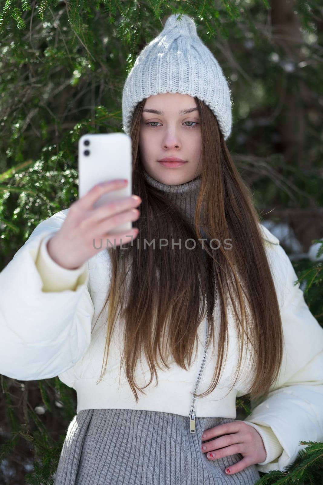 Teen girl 17 year old using cell phone in vertical orientation, shooting video to send to social networks through mobile application. Female wearing hat, puffer bomber jacket, standing in pine forest