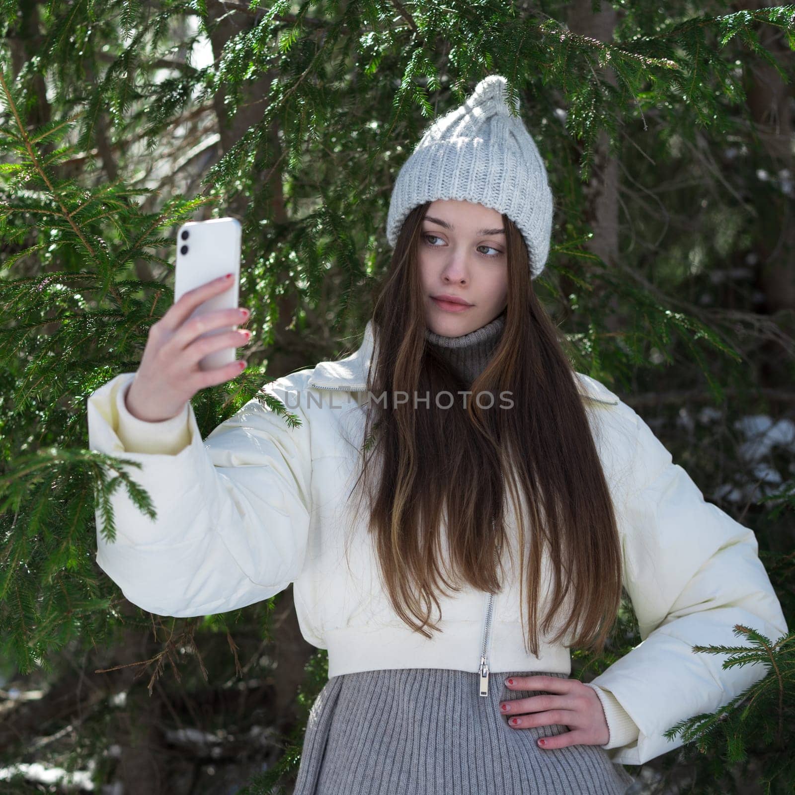Portrait of Caucasian teen girl using mobile phone making pictures selfie to capturing moments from pine forest. Serious teen model with long hair, no make-up wearing knitted hat, puffer down jacket