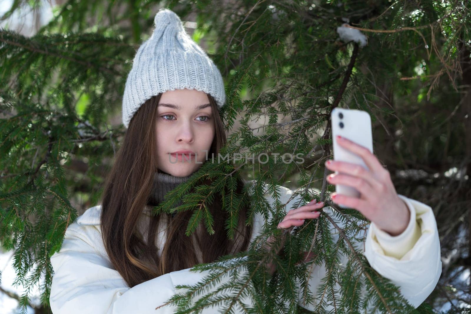 Portrait teenager using cell phone on pine forest, holding mobile phone in hand and looking at phone camera, posing, making video selfie. 17 year old female model in knitted hat, puffer bomber jacket