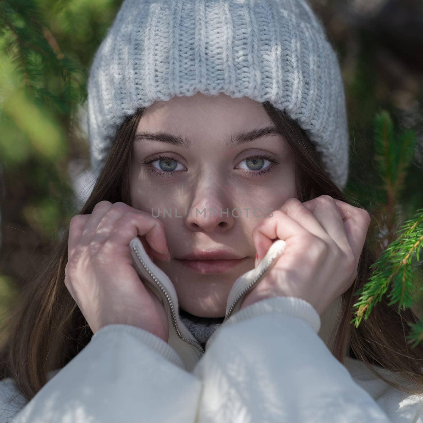 Cropped shot of teenager holding collar of bomber jacket with his hands in cold winter weather in forest, looking at camera. 17 year old model with gray eyes, no make-up wearing in knit hat, jacket