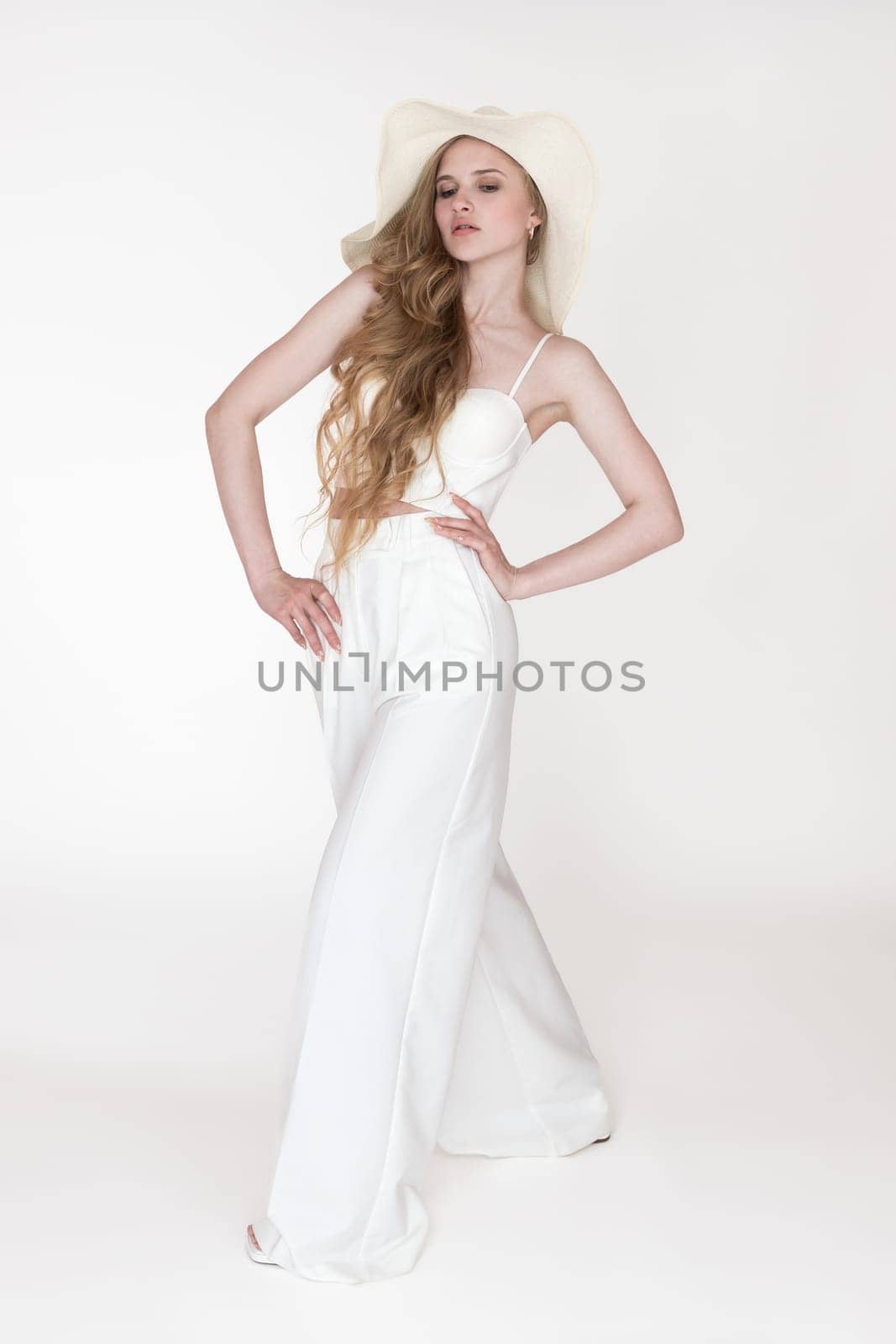 Full length portrait of young blondie fashion model 21 year old with wavy long hair in straw hat, white sculpting cupped corset top and pants standing on white background. Caucasian woman looking down
