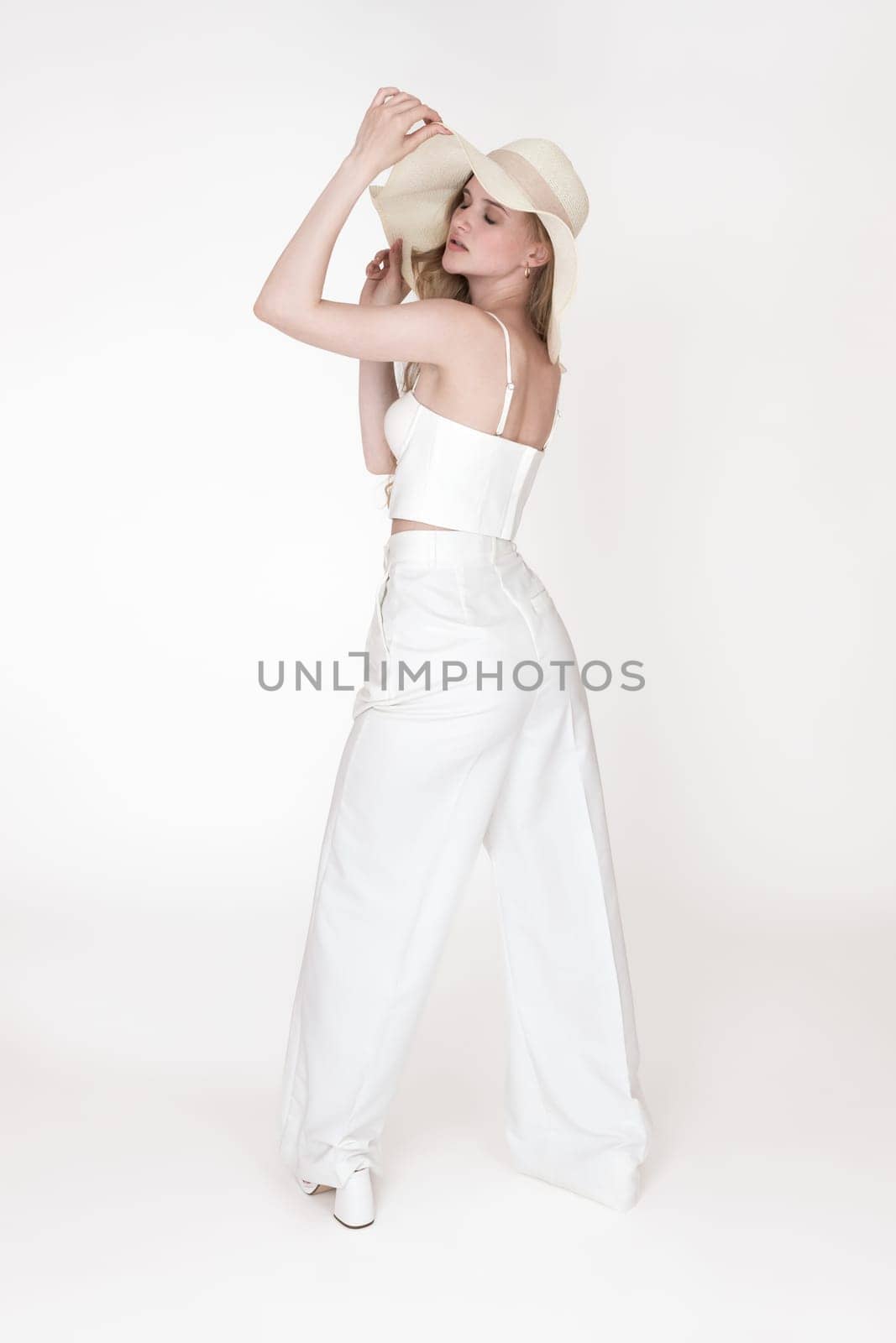 Back view of woman with eyes closed holding brim of sun hat with both hands. Fashionable model well dressed in straw hat, white sculpting cupped corset top and trousers, standing on white background