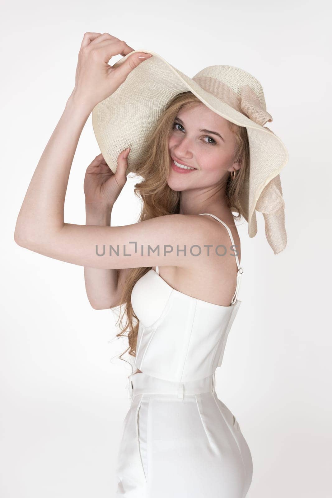 Smiling blonde fashion model looking over shoulder, raised hands holding brim of hat, well dressed in white floppy sun hat, cupped corset top, trousers. Portrait of happiness woman on white background