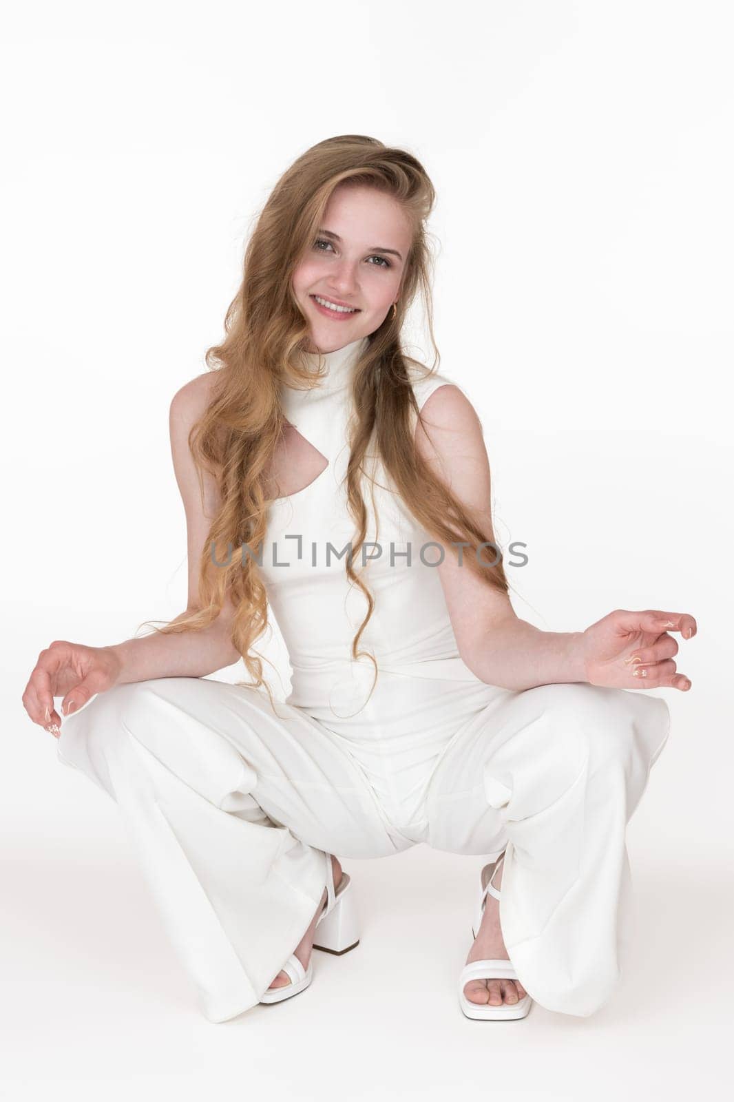 Beautiful blonde woman dressed in white jumpsuit sitting squatting position and looking at camera by Alexander-Piragis