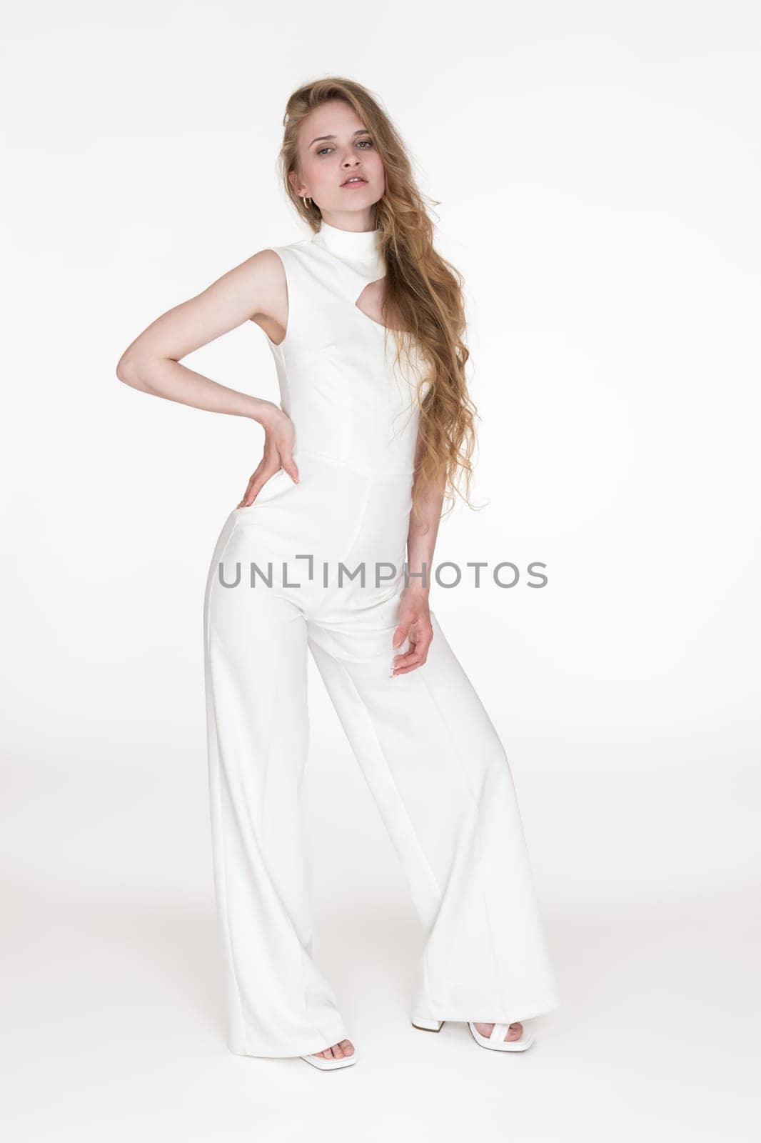 Young woman wearing white long jumpsuit office looks looking at camera, standing on white background by Alexander-Piragis