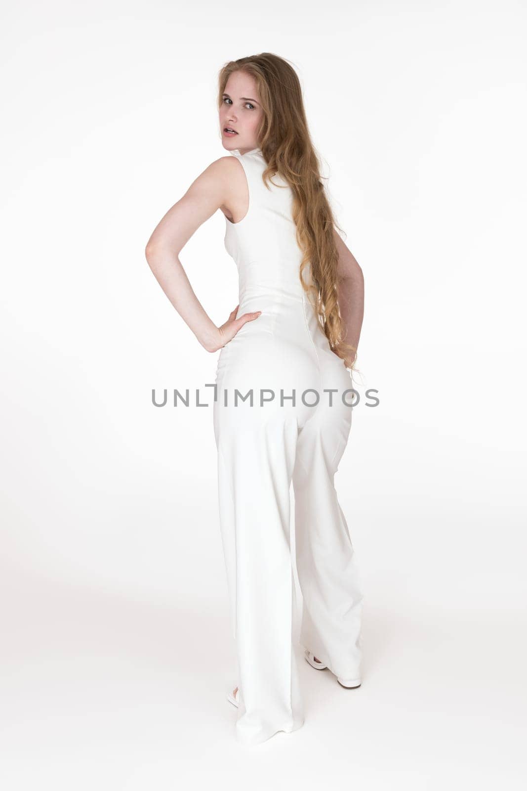 Back view of young adult woman looking over shoulder at camera dressed in white long jumpsuit, standing arms akimbo on white background. Beautiful sexy woman model with long blonde hair. Studio shot