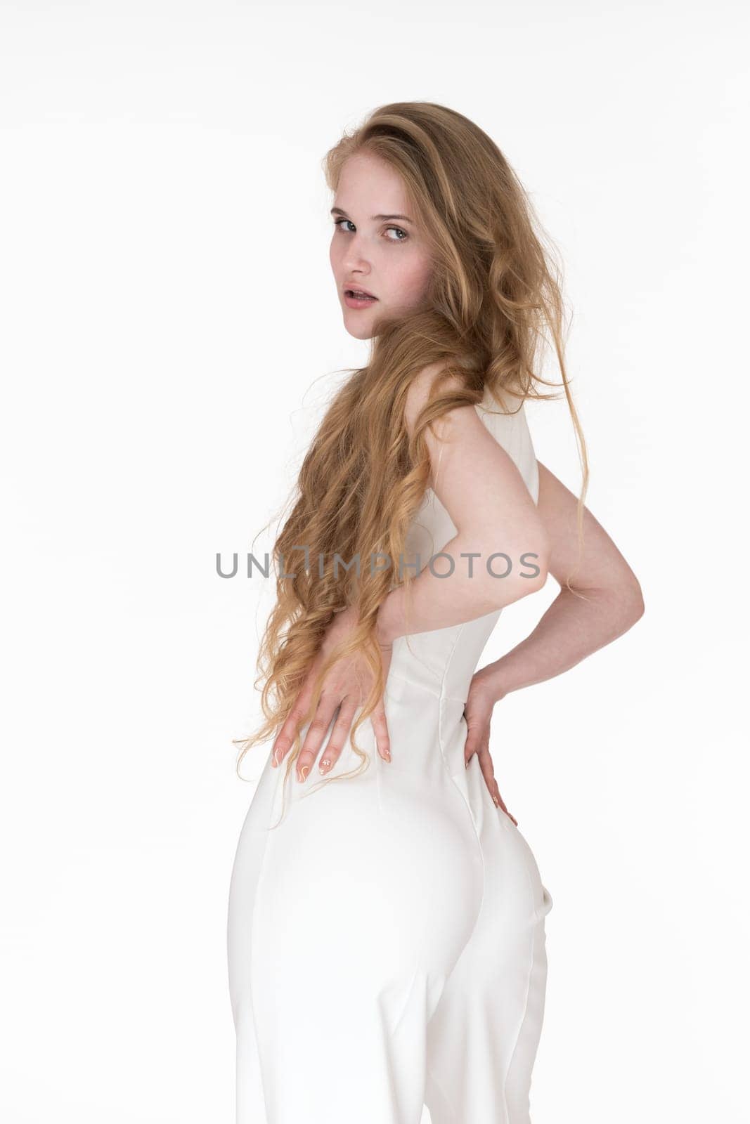 Back view of young blonde woman looking over shoulder at camera. Caucasian model with blonde long wavy hair dressed in white jumpsuit. Waist up female arms akimbo on white background. Studio shot