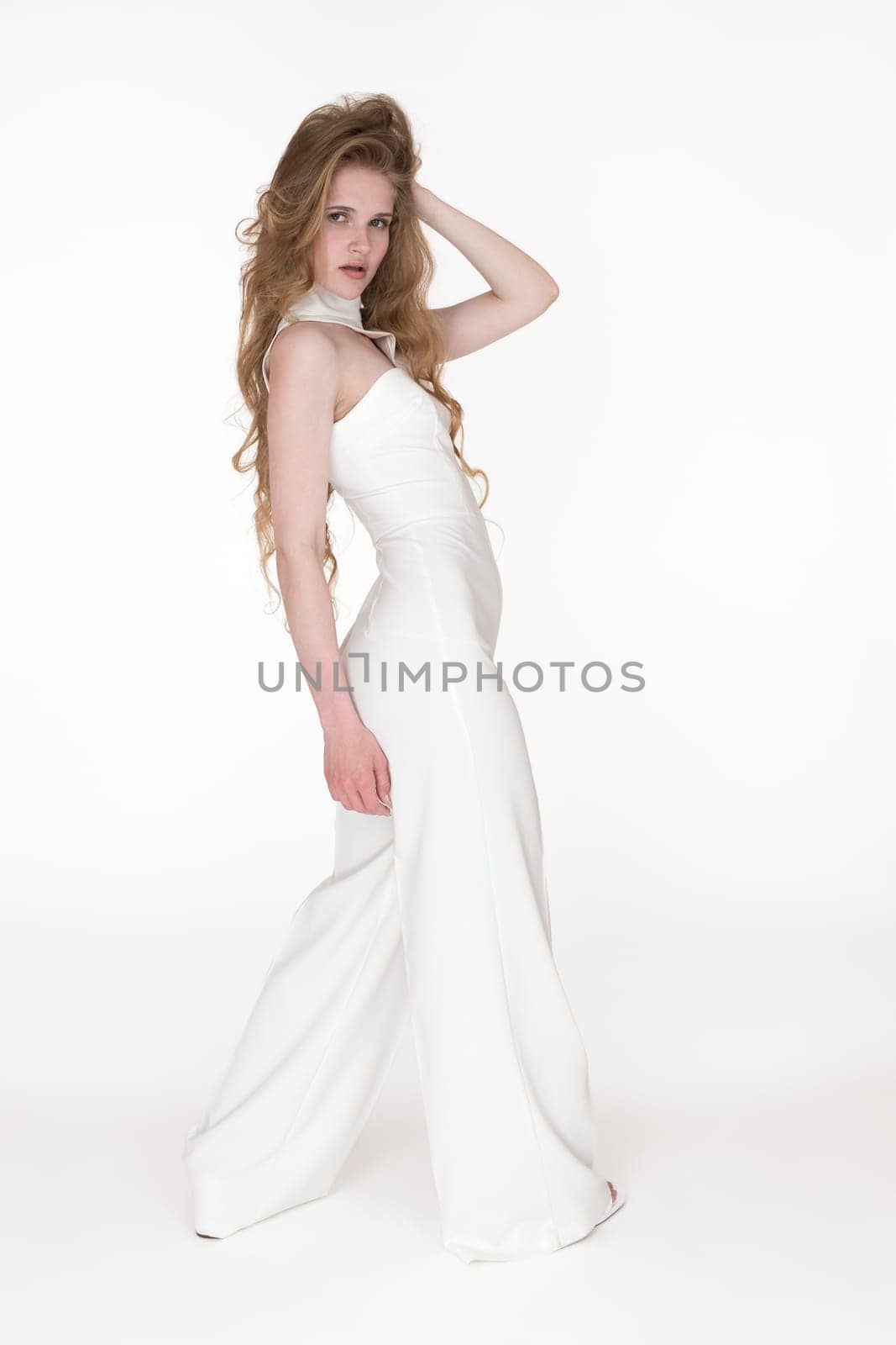 Young woman with hand behind head looking over shoulder at camera dressed in white long jumpsuit on white background. Full shot of sensuality Caucasian fashion model with long blonde hair 21 years old