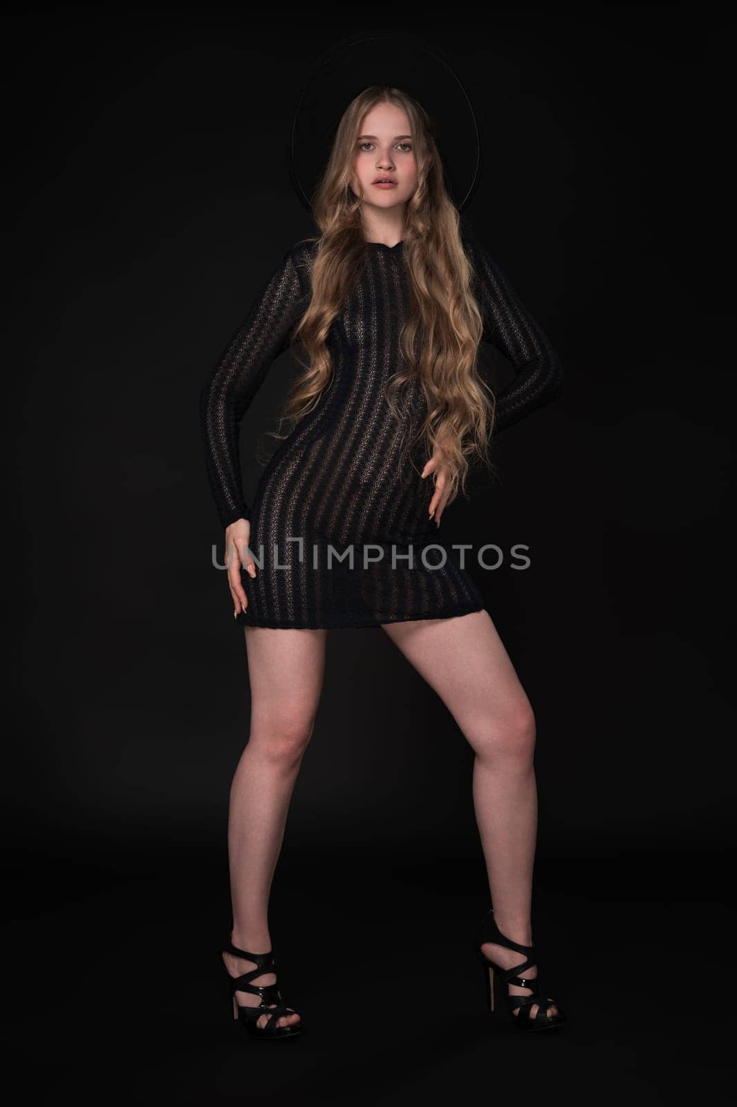 Woman in full length exudes confidence and poise. Her long legs are accentuated as she strikes pose on black background. Model wears black short knitted tight dress, felt hat and high-heeled shoes