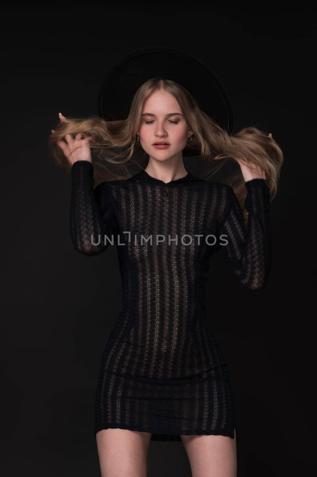 Portrait slim young blonde woman with closed eyes and hands in motion straightening long hair. Model in short tight knitted translucent dress and wide-brimmed felt hat. Black background adds to drama