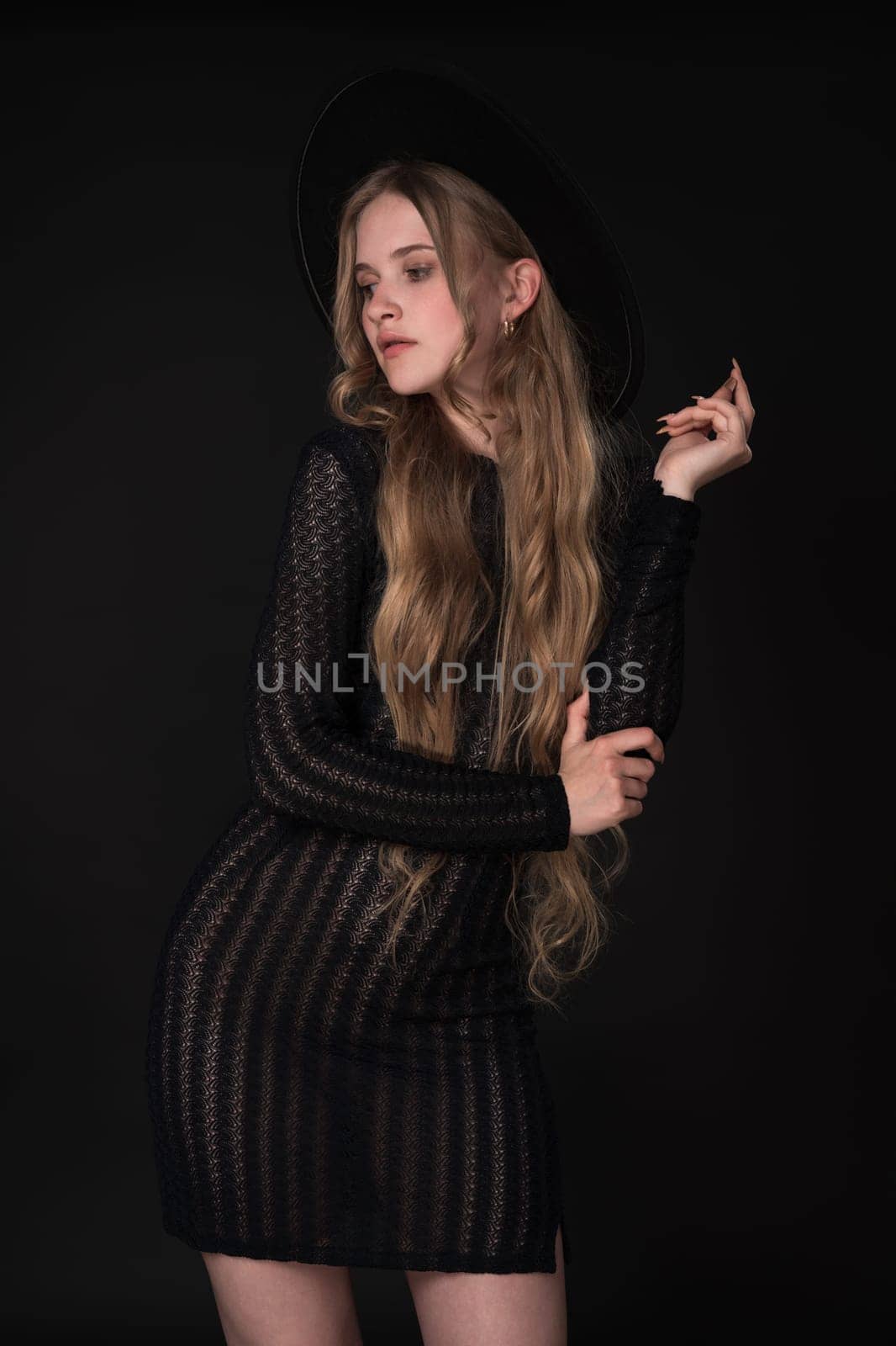 Portrait of shy young blonde looking away, shyly raising one arm bent at elbow, supporting it with other. Blonde with long hair wears short black tight knitted see-through dress, wide brimmed felt hat