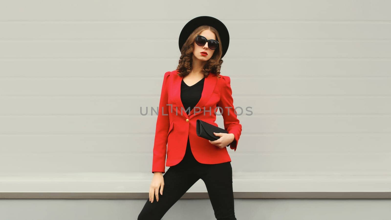 Stylish elegant woman posing in business suit, red blazer jacket, black round hat with handbag clutch
