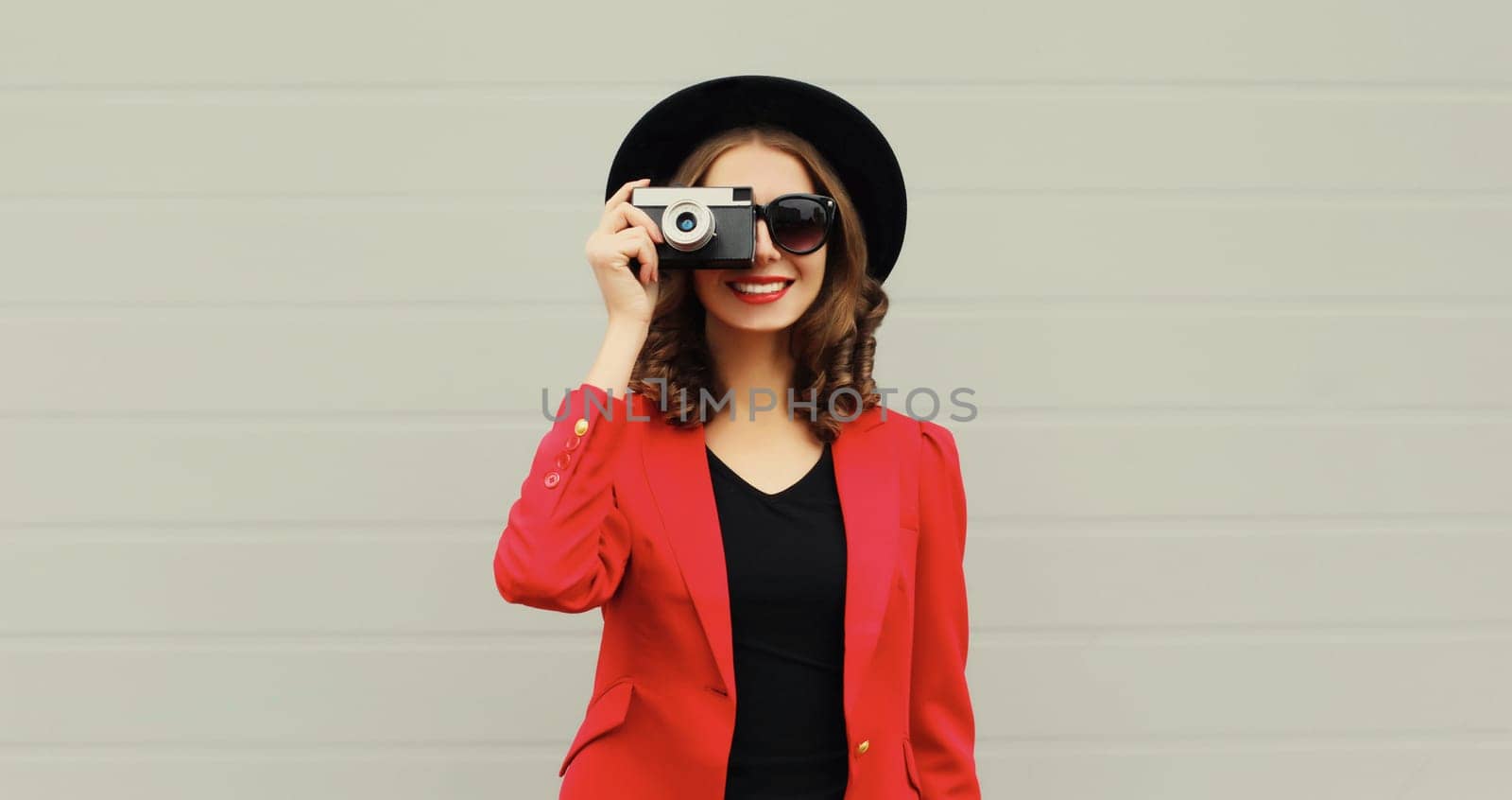 Beautiful stylish young woman photographer with film camera taking picture in black round hat, red blazer jacket