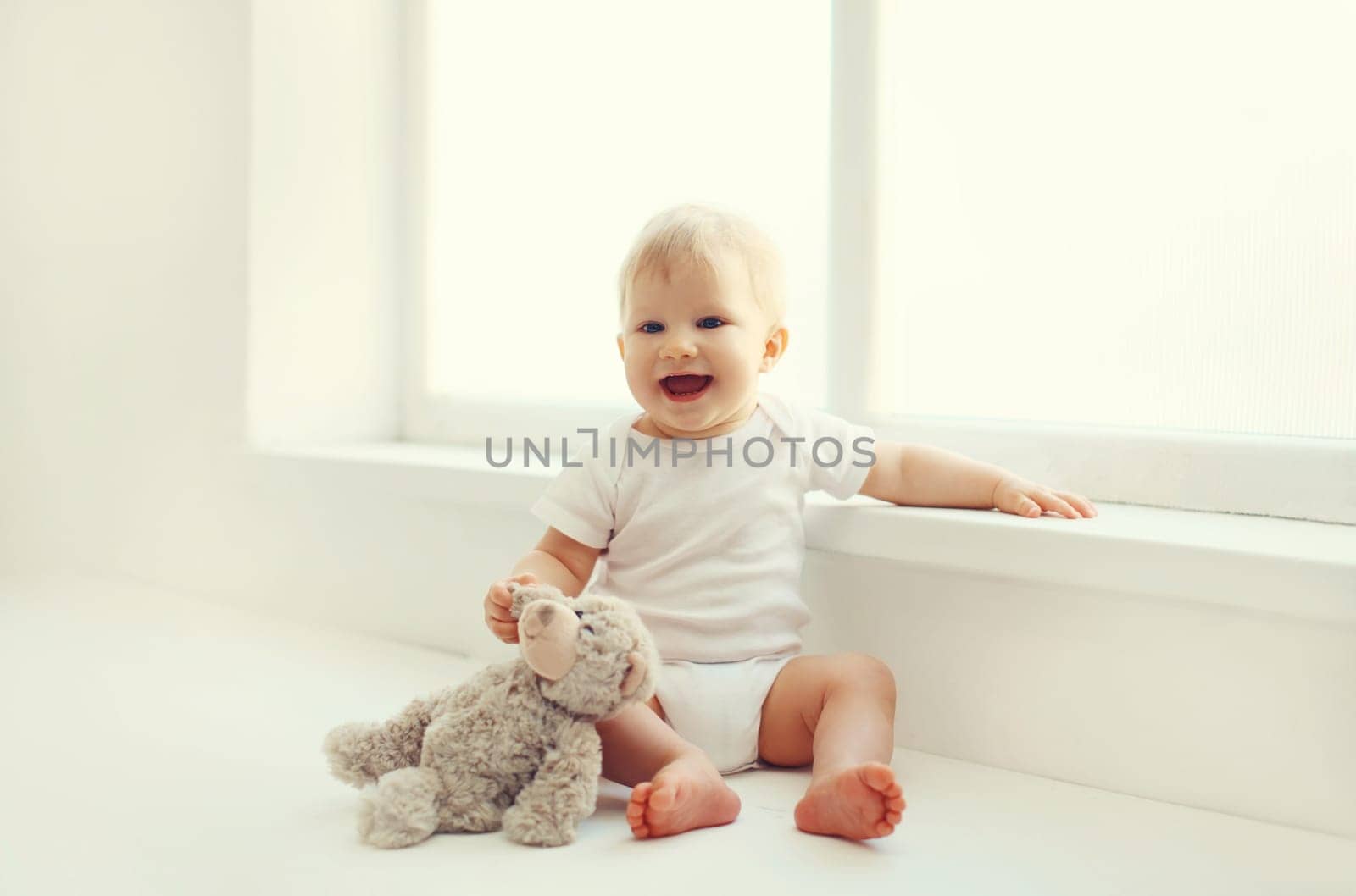 Happy cute little baby playing with teddy bear toy on the floor in white room at home