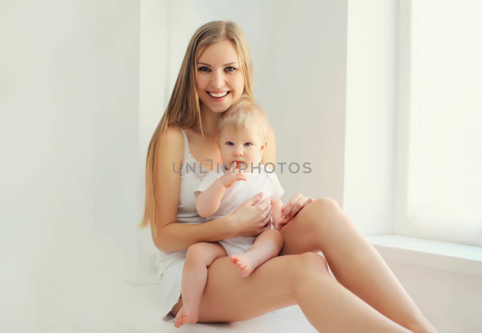 Happy smiling young mother playing with cute baby sitting together in white room at home