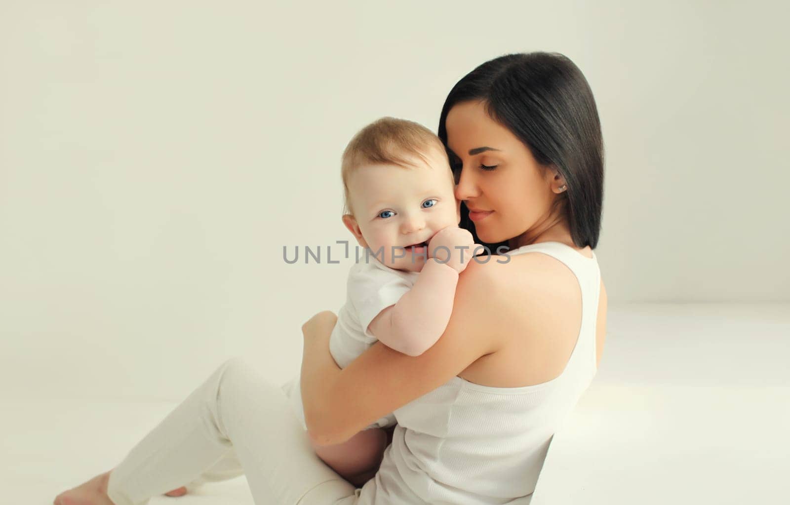 Happy young mother holding her cute baby sitting together in white room at home
