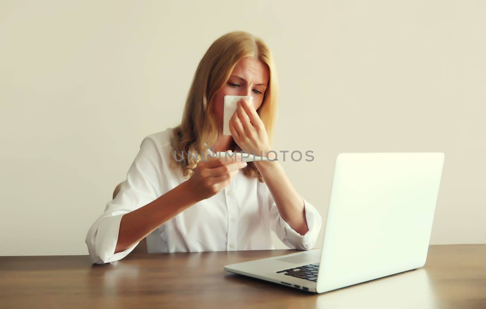 Sick woman sneezing blow nose using tissue, measuring checking body temperature with thermometer working with laptop sitting at desk in office