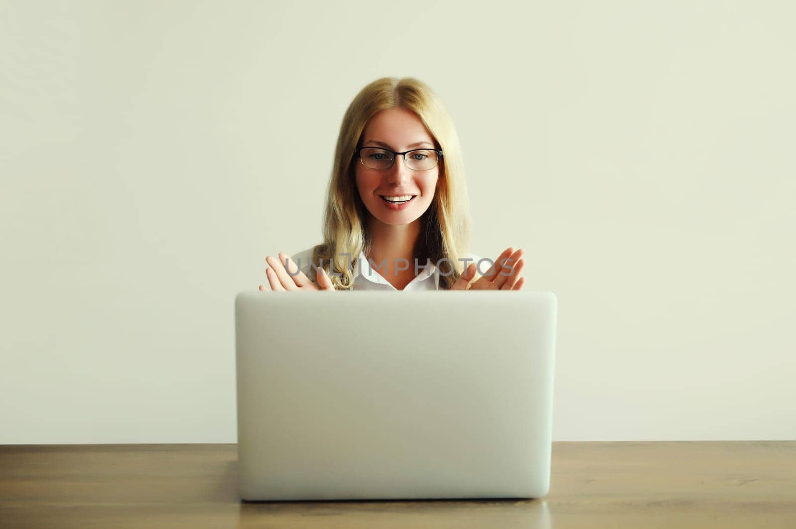 Happy young woman working on laptop greets communicates with friends via video call sitting at desk in office or home